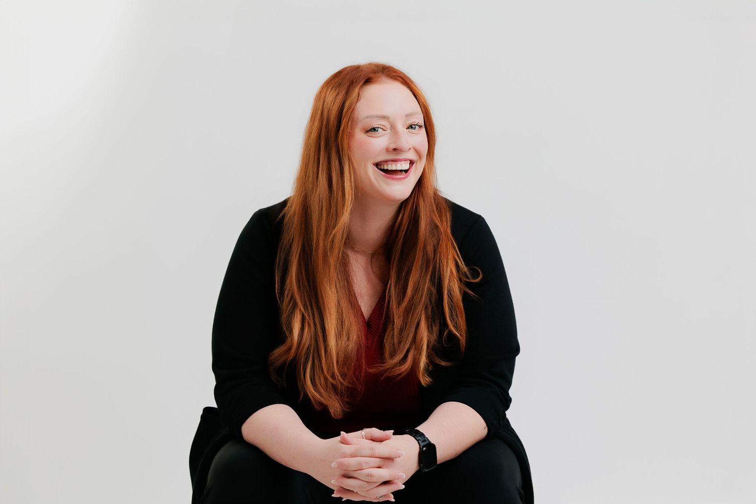 A professional woman in black clothing sits against a white backdrop, laughing with bright red hair falling past her shoulders.