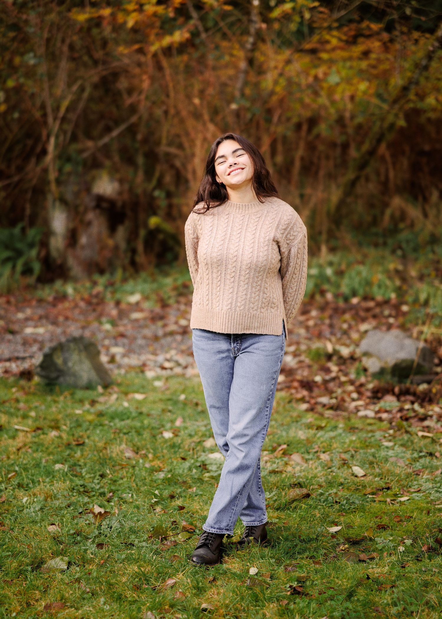 A person in a cozy beige sweater and light blue jeans poses playfully on grass covered in autumn leaves.