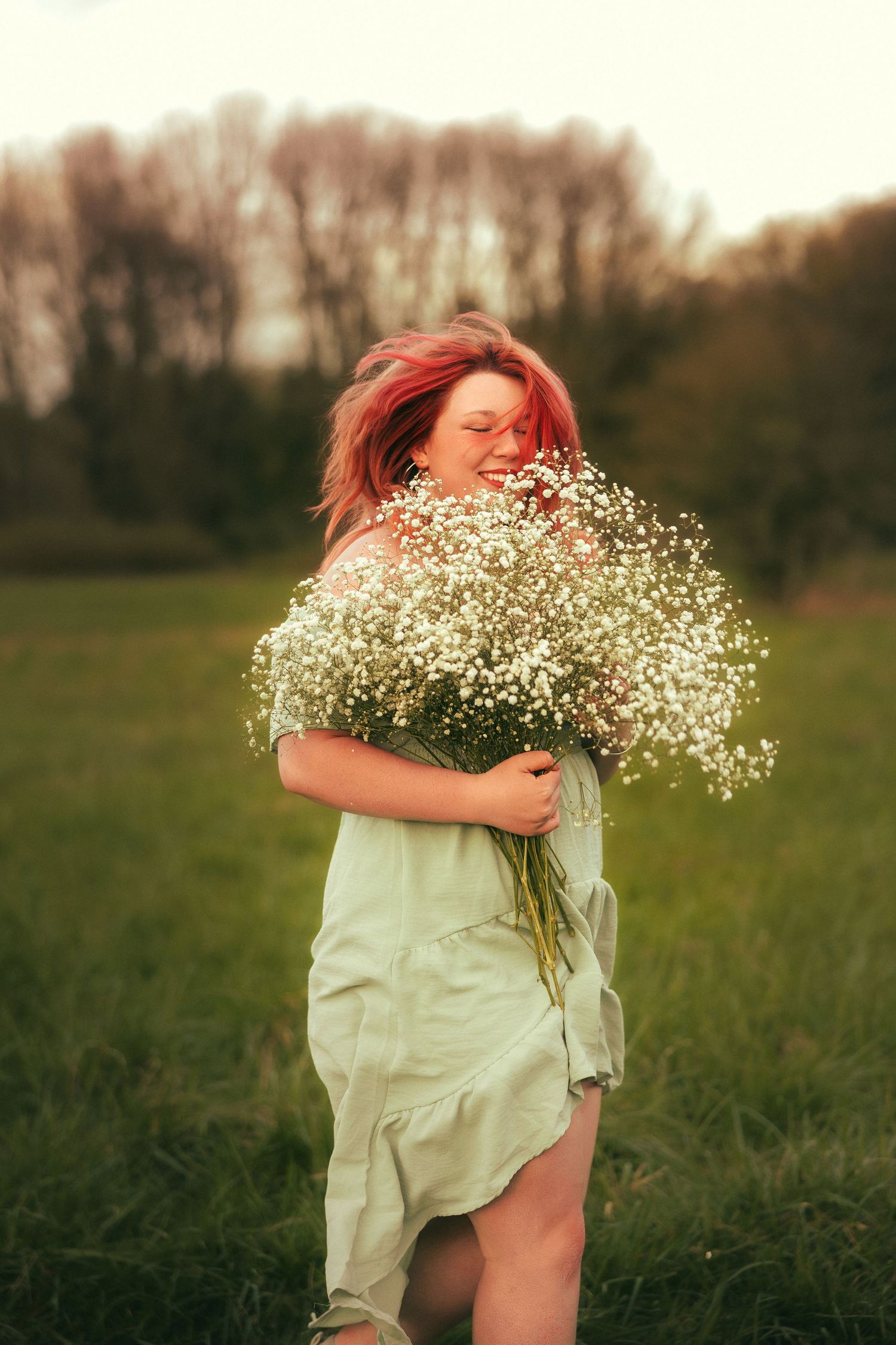 A figure in a flowing white dress holds a large bouquet of baby's breath flowers in a sunlit meadow at dusk.
