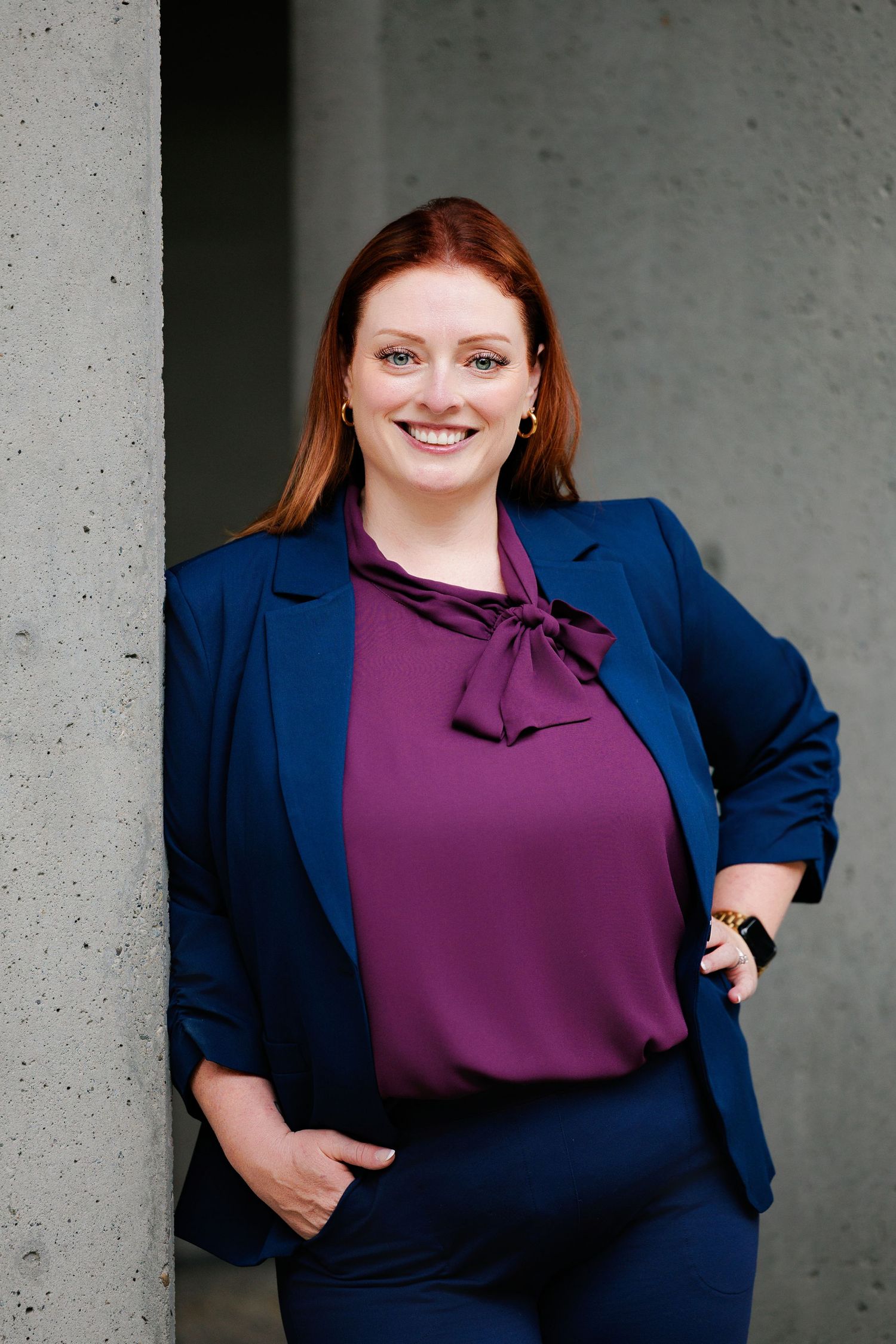 Professional portrait of someone wearing a navy blazer and purple tie-neck blouse against a concrete wall background.
