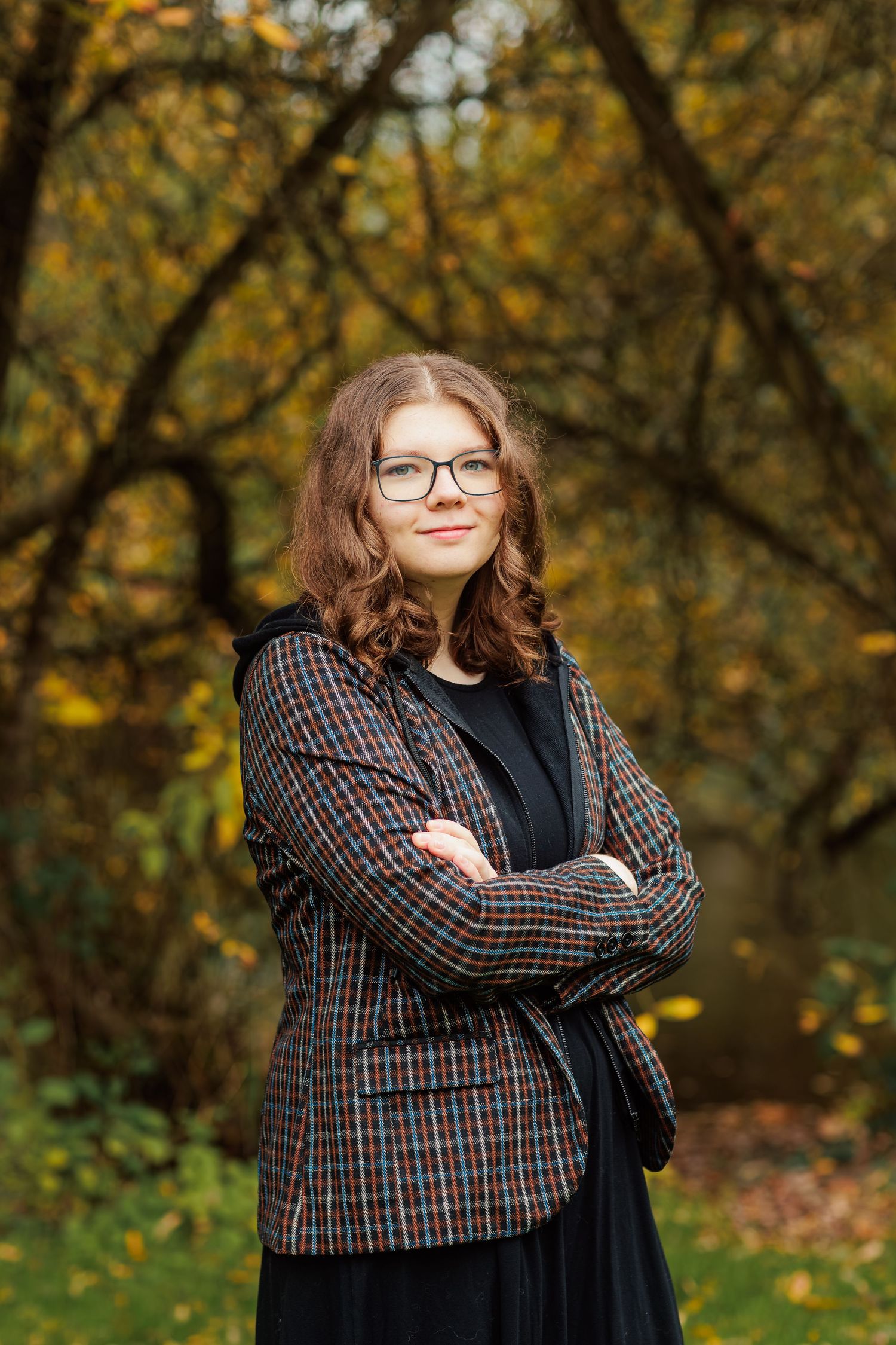 A person in a plaid blazer and black shirt poses confidently against an autumn backdrop with golden foliage.