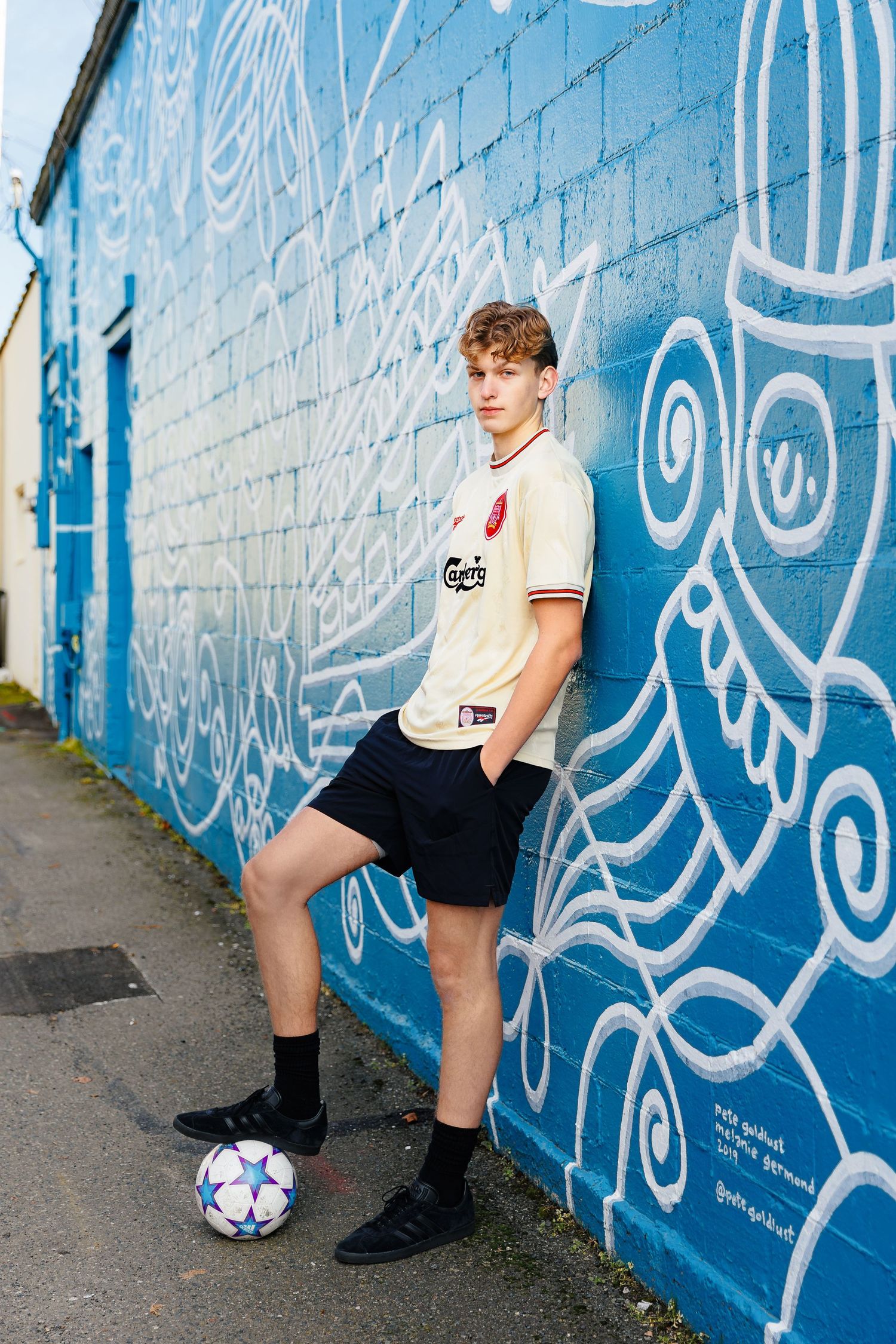 Soccer player in white jersey poses against blue graffiti wall with ball in urban setting.