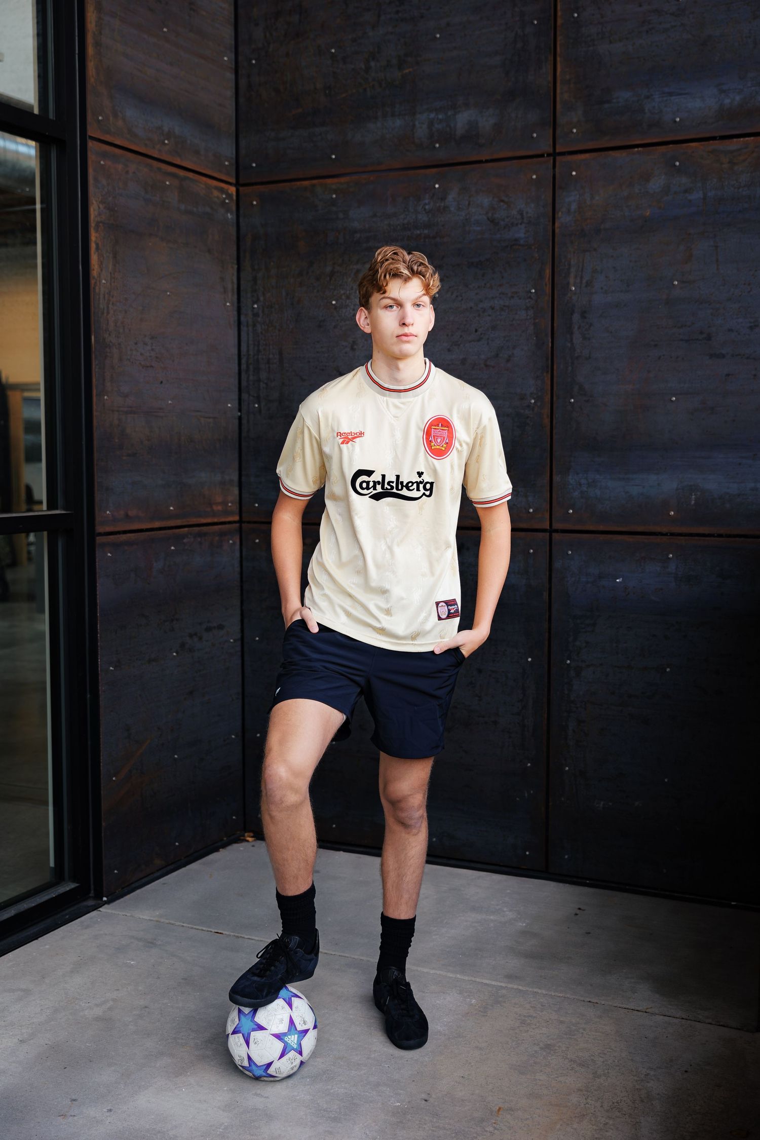 Soccer player wearing a vintage Carlsberg jersey poses with a ball against dark metal wall.