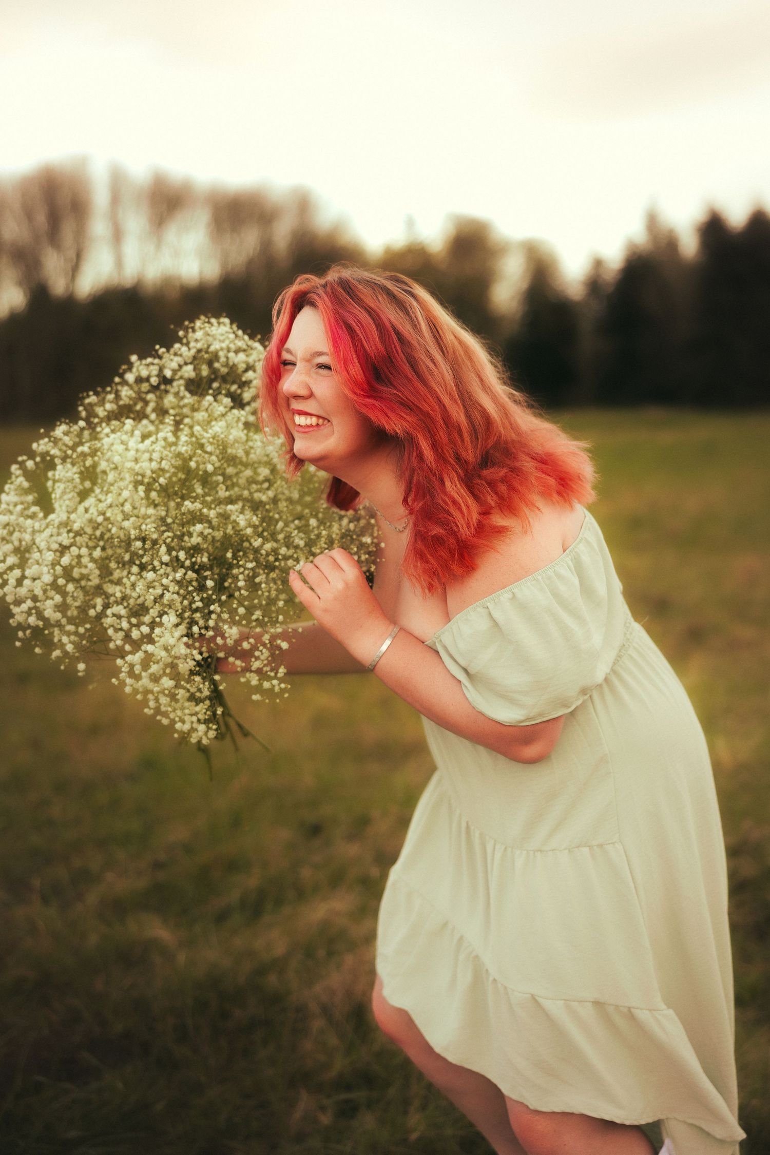 A figure in a white dress holds baby's breath flowers while smiling in a sunlit meadow at golden hour.