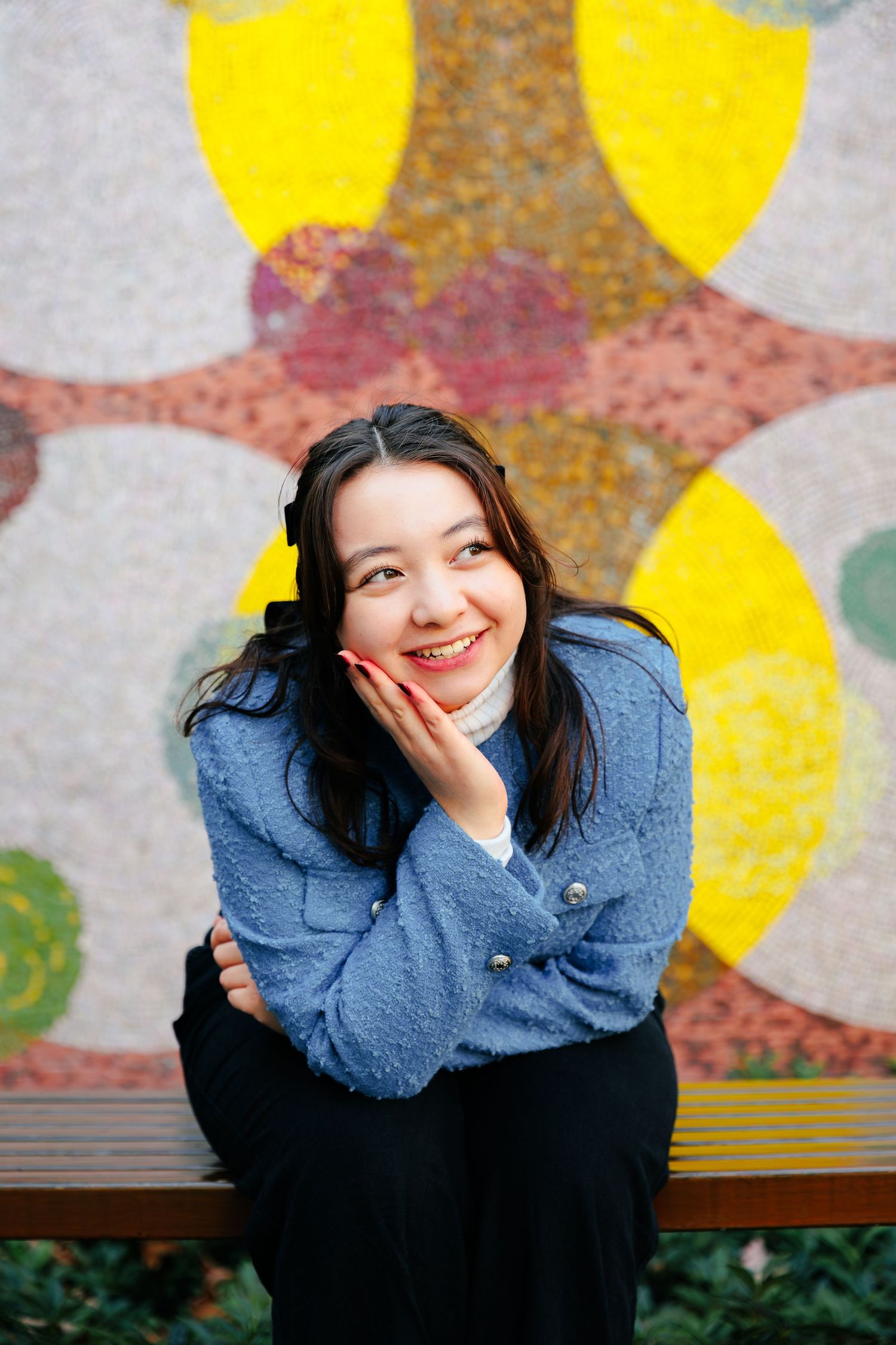 A person in a blue sweater poses cheerfully against a colorful wall with yellow circles and abstract patterns.