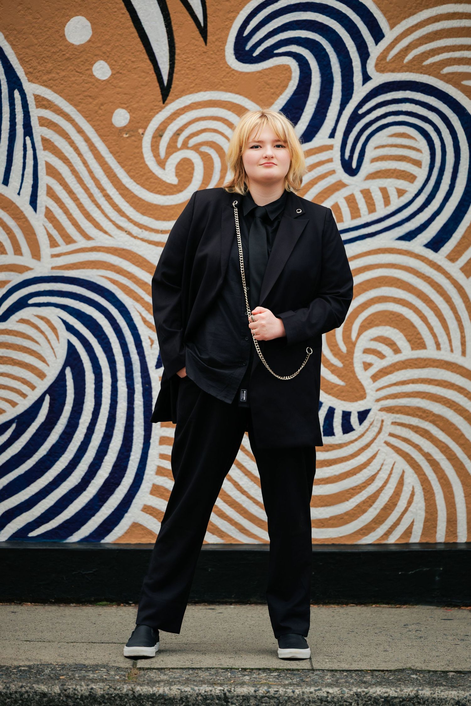 Person in all black outfit poses against wall with navy blue and white wave pattern mural in urban setting.