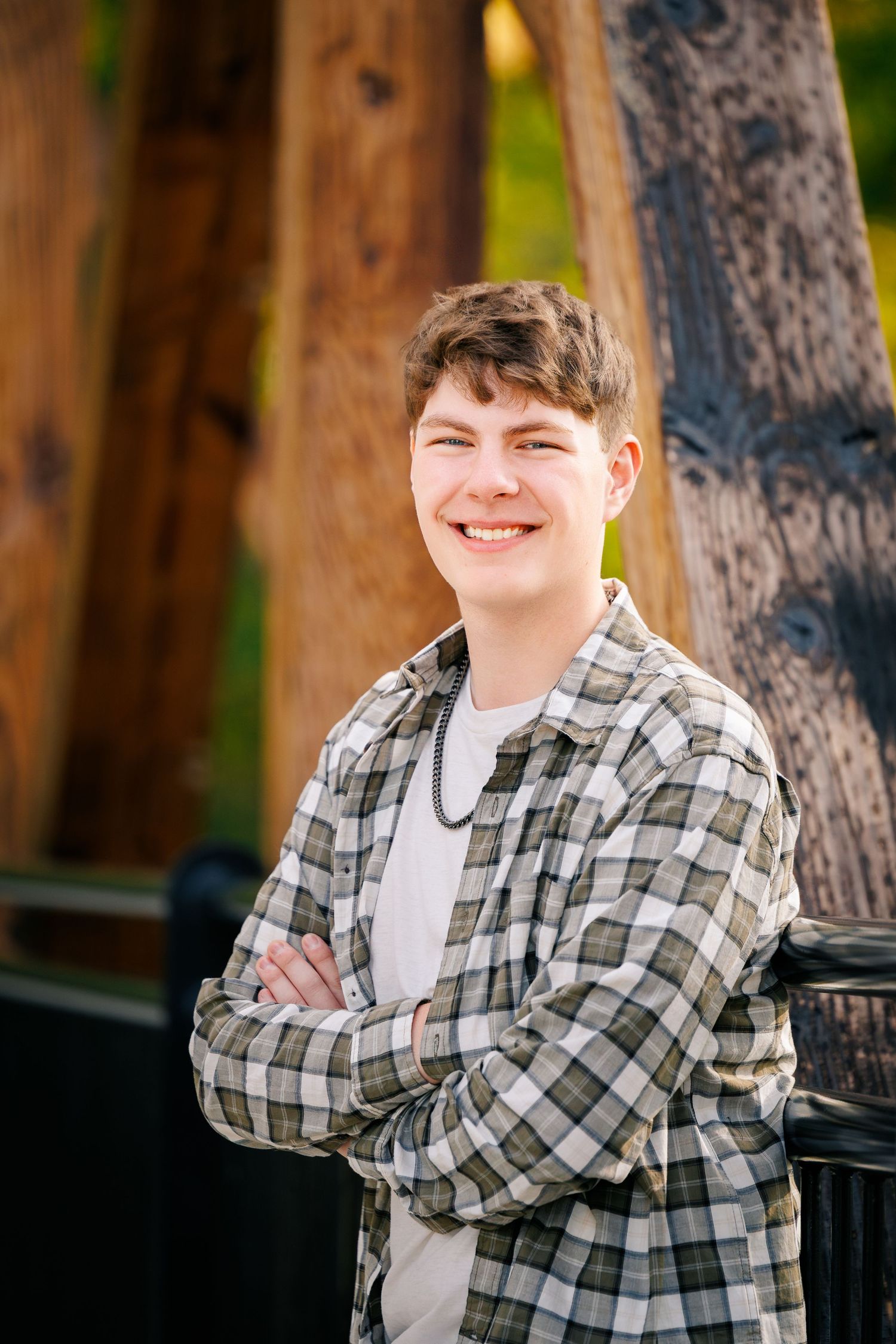 A young person in a plaid flannel shirt and white t-shirt smiles while standing outdoors near wooden posts.