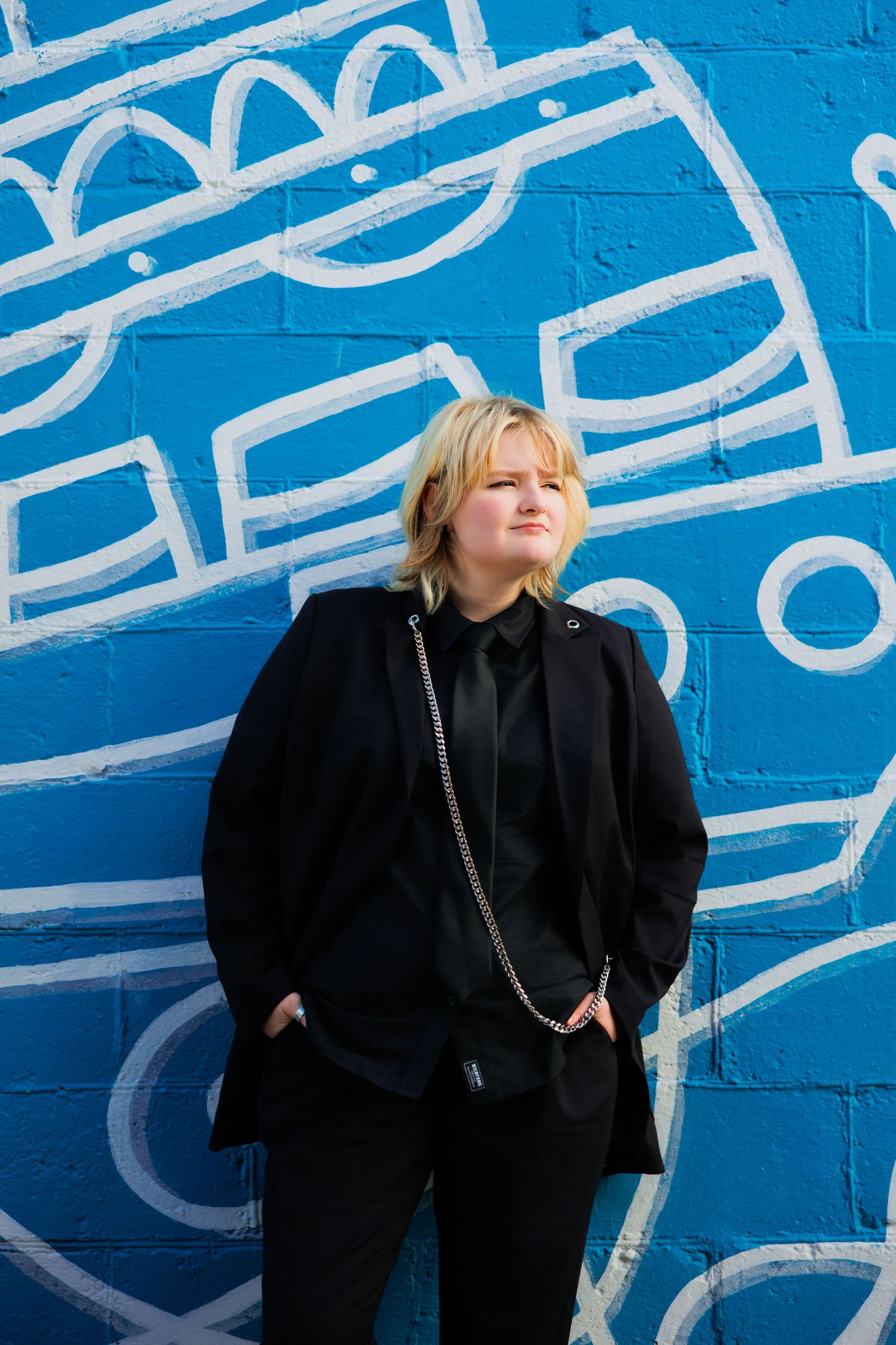 Figure in black outfit poses against vibrant blue wall with white graffiti art pattern in artistic portrait composition.