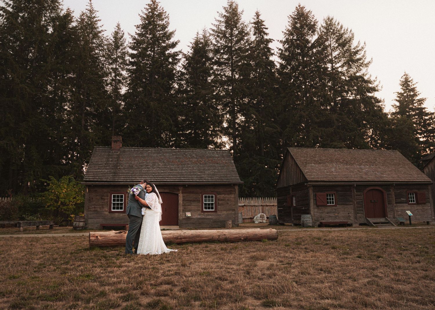 A multiracial bride and groom embrace in front of historic rustic buildings at sunset