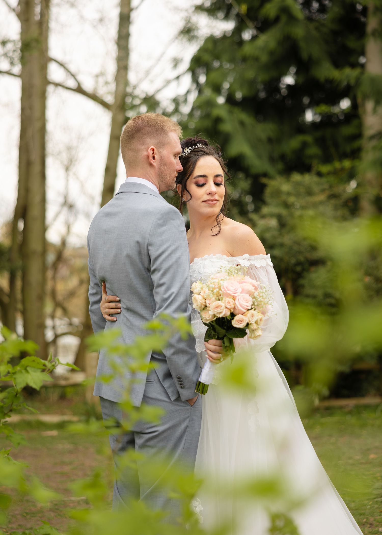 A romantic wedding moment captured in a wooded outdoor setting with soft natural lighting and pastel flowers.