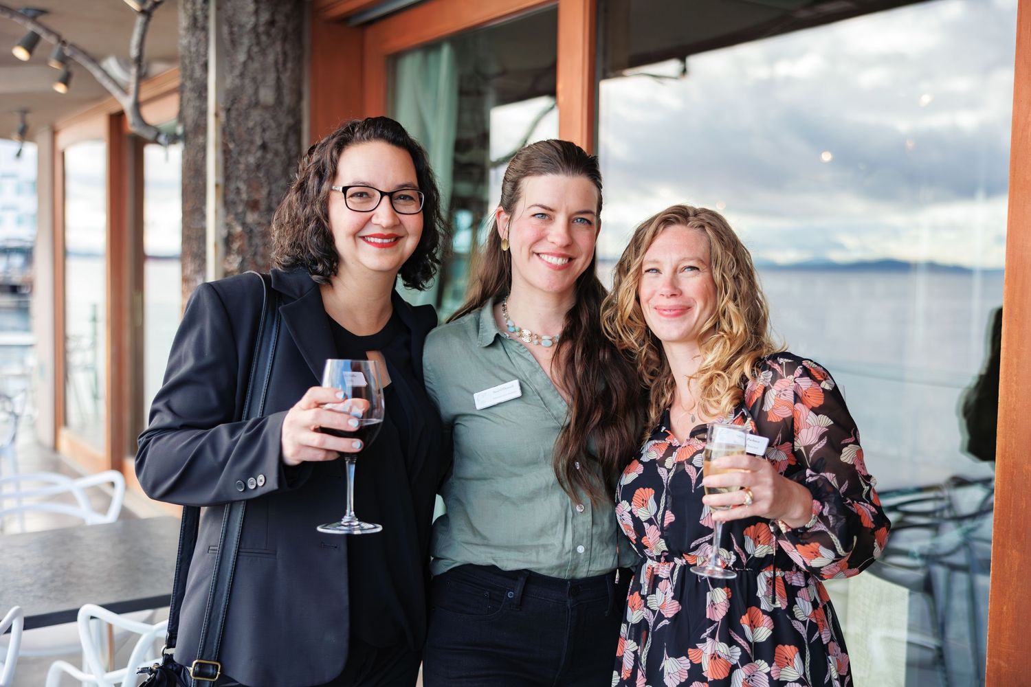 Three work friends enjoying drinks and conversation on an outdoor patio with water views.