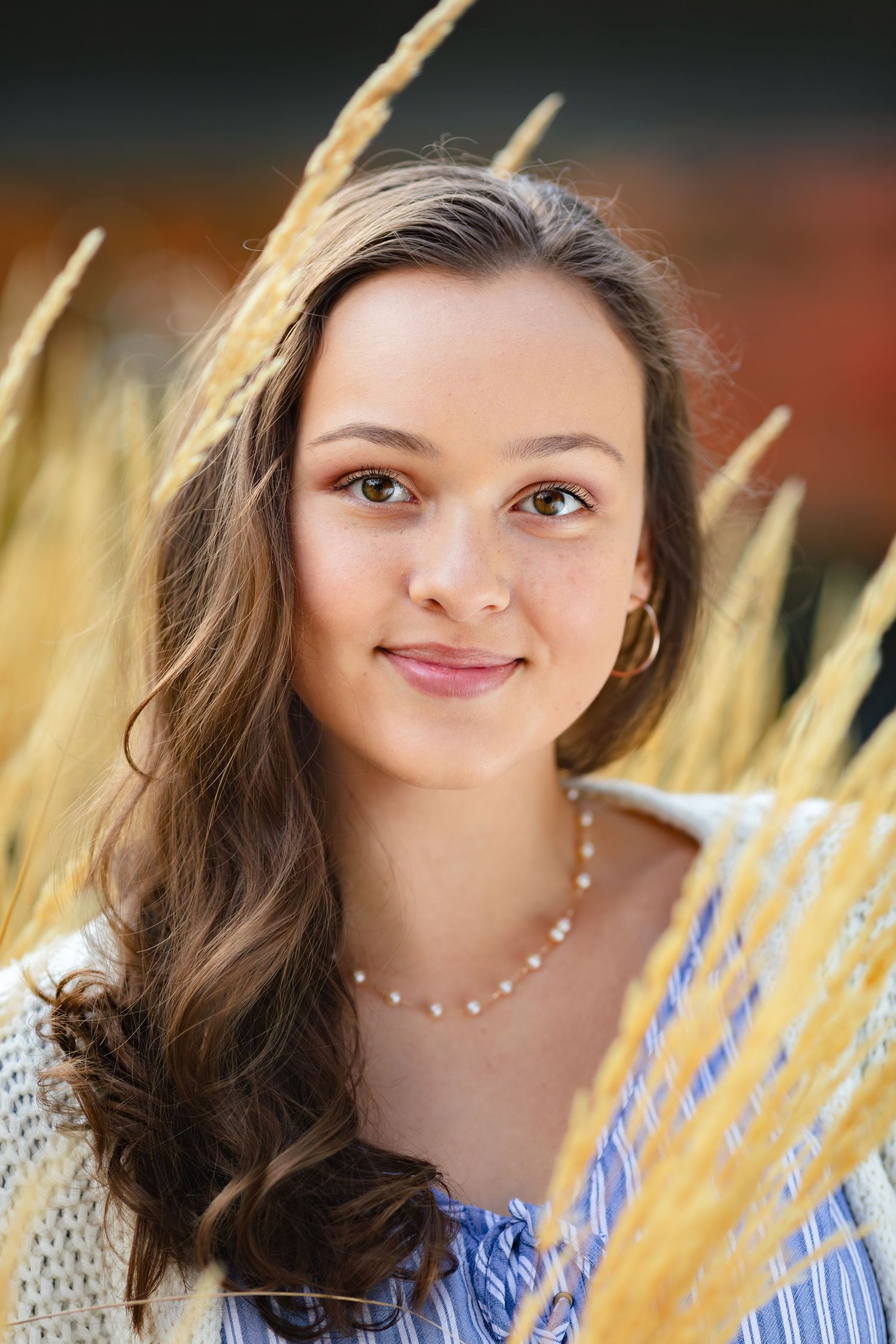 Portrait composition with long brown wavy hair and pearl earrings against warm golden lighting and blurred background.