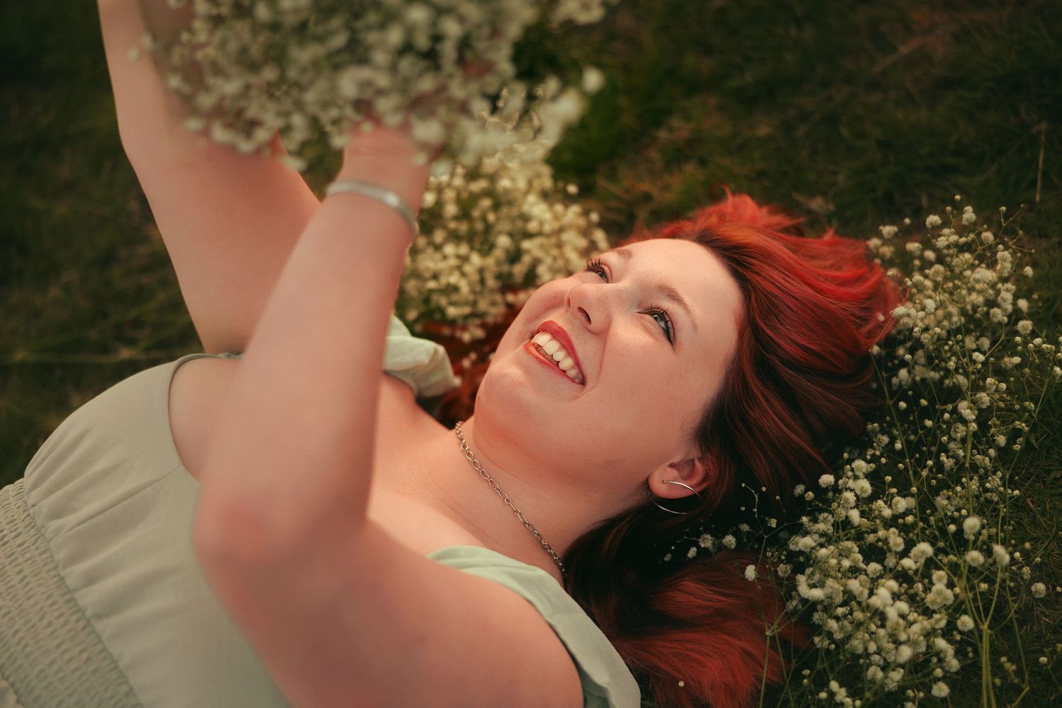 A teenaged girl with vibrant red hair lies in a field of grass holding a baby's breath bouquet, smiling in the warm sunlight.