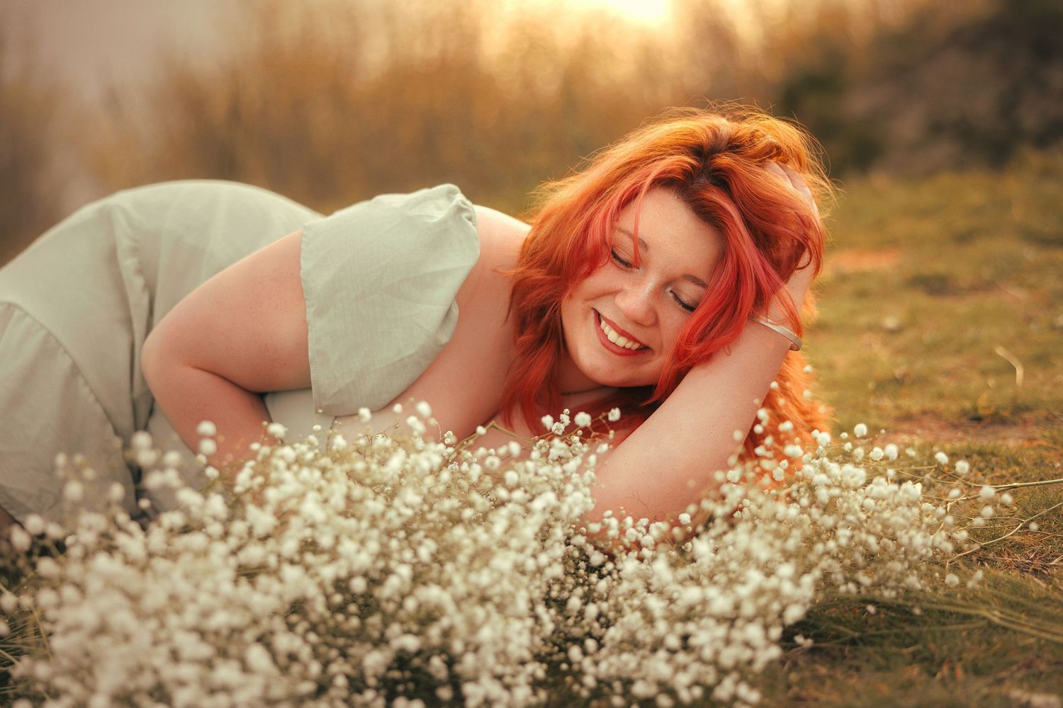 A teen girl with vibrant red hair lies in a field of grass with baby's breath during golden hour.