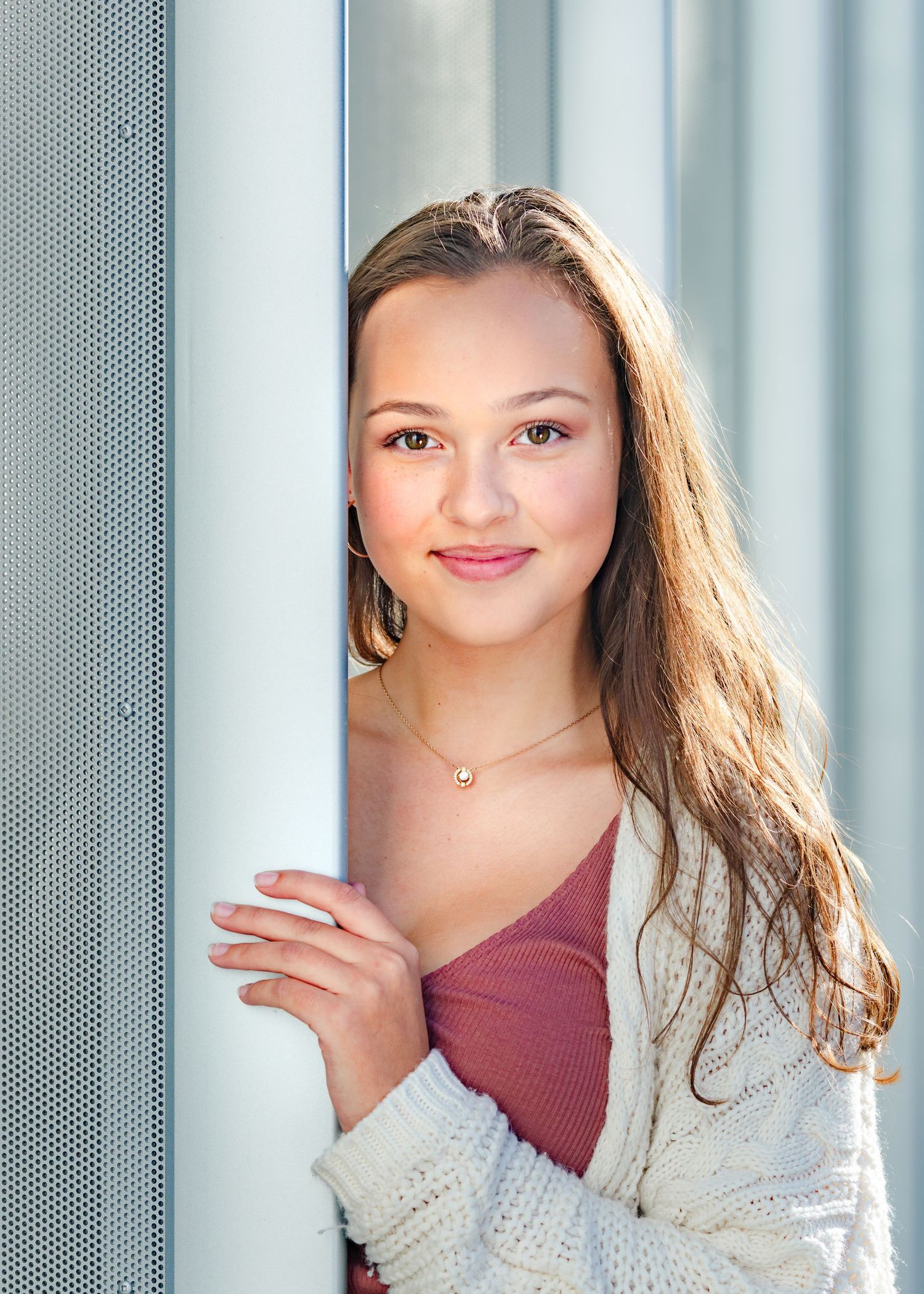 Young woman in burgundy top and white cardigan poses against light curtain background with natural lighting.