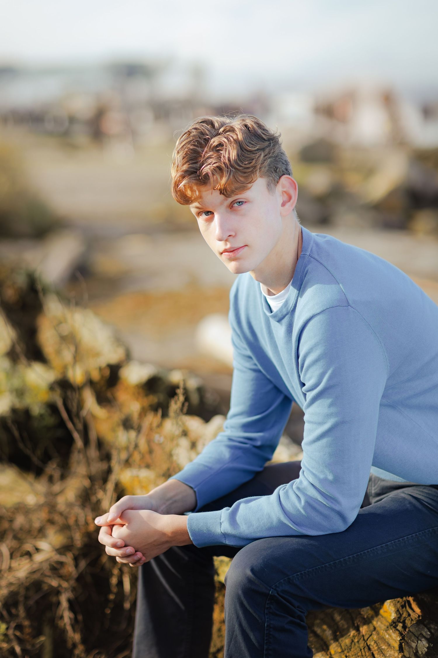 A high school senior in a light blue sweater sits thoughtfully on a driftwood log on the beach at golden hour.