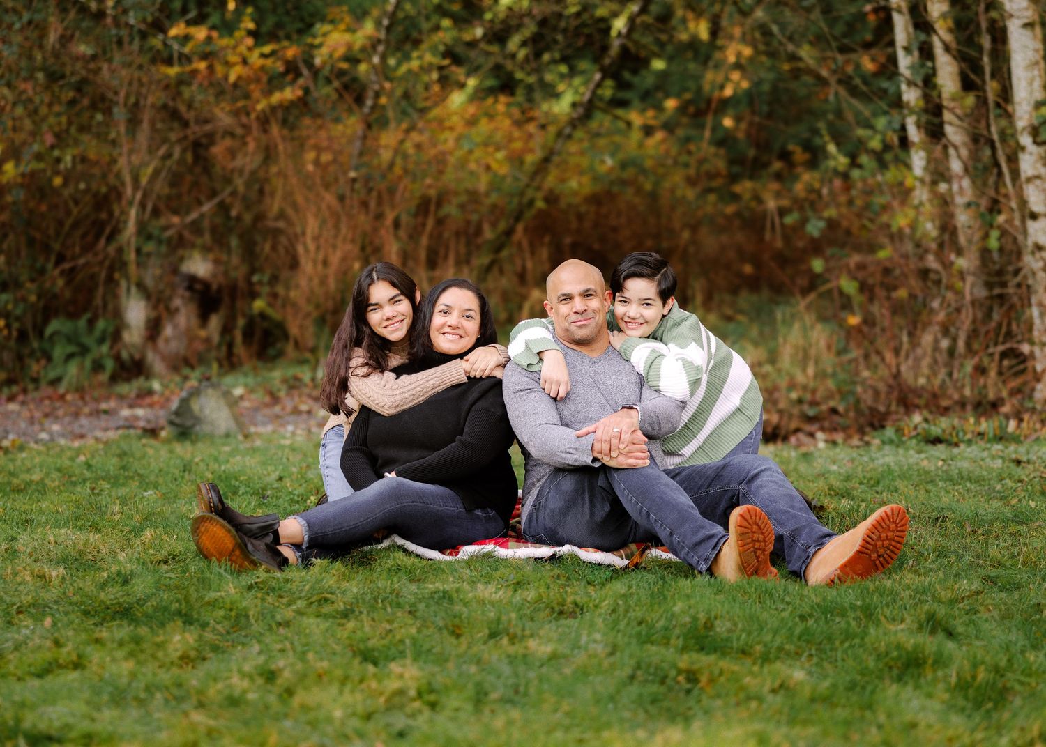 A family sits together on a blanket enjoying a warm autumn day in a park setting with fall foliage in the background.