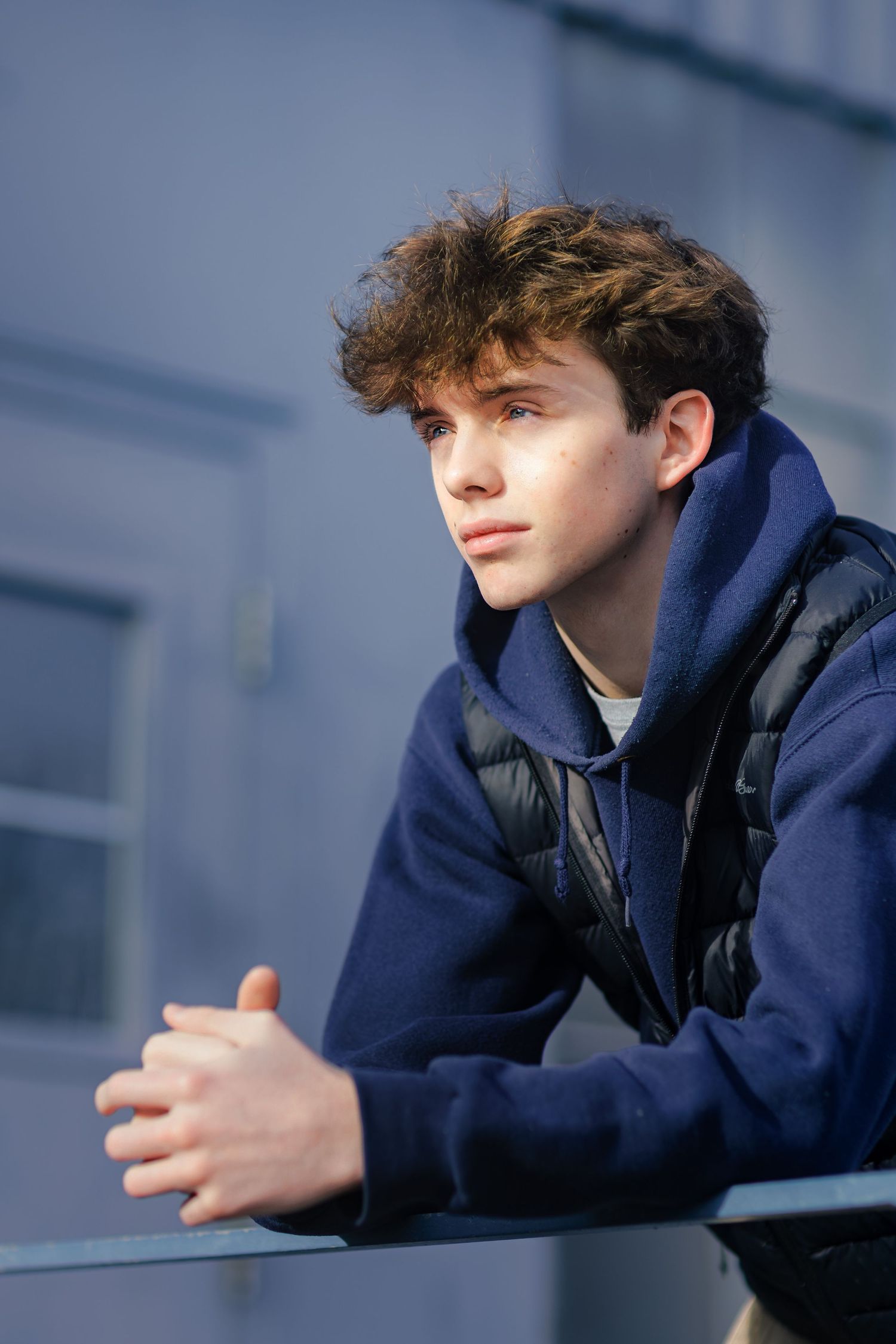 A person in a navy hoodie and black vest leans thoughtfully against a railing while looking into the distance.