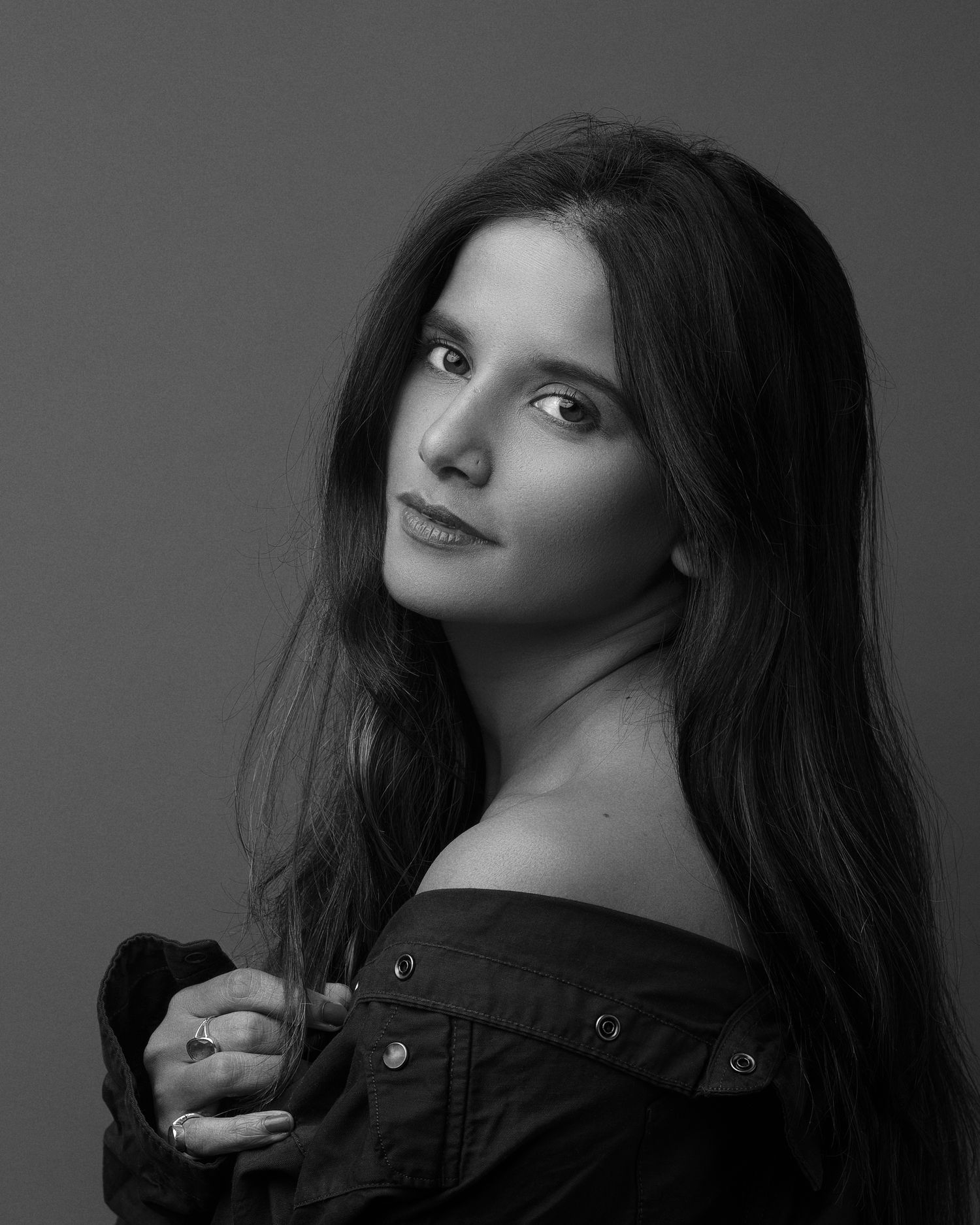 Black and white portrait of a young woman with long dark hair wearing an off-shoulder top against a dark background.