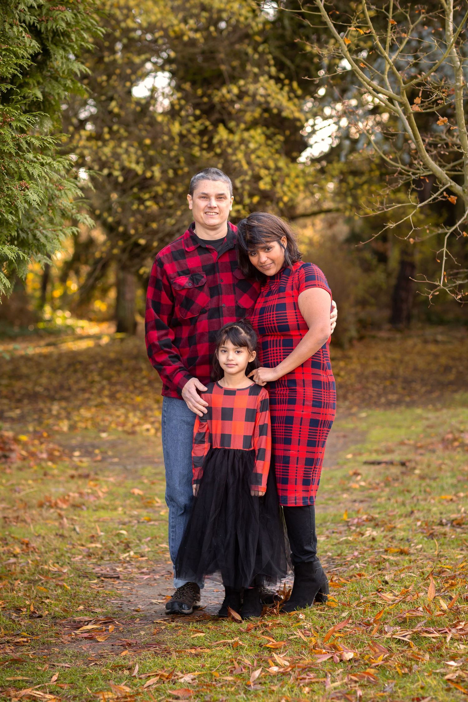 Family wearing coordinating red plaid outfits poses together on an autumn path with fallen leaves and trees.