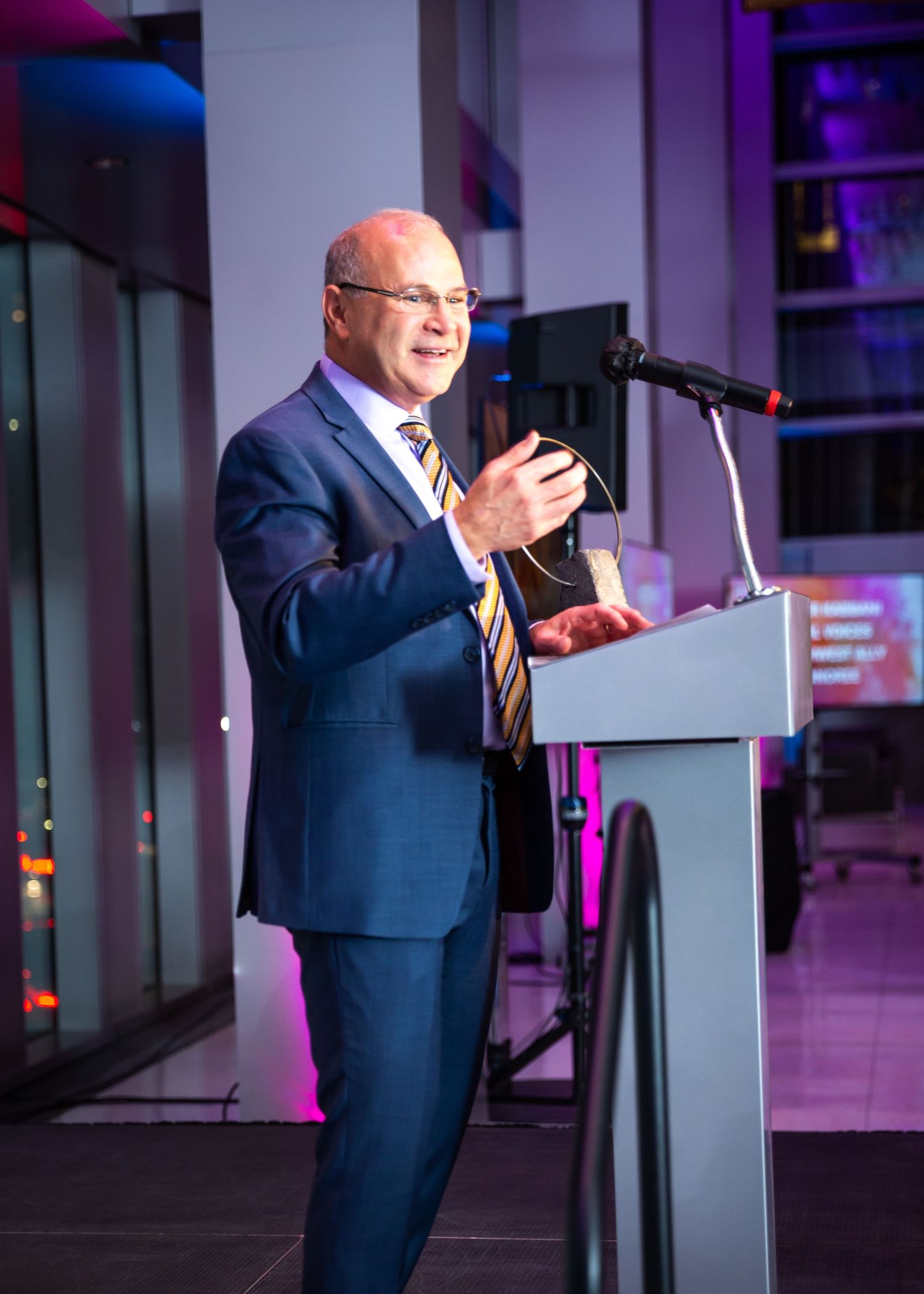 A speaker in a navy blue suit gestures while presenting at a podium with dramatic purple and blue lighting.