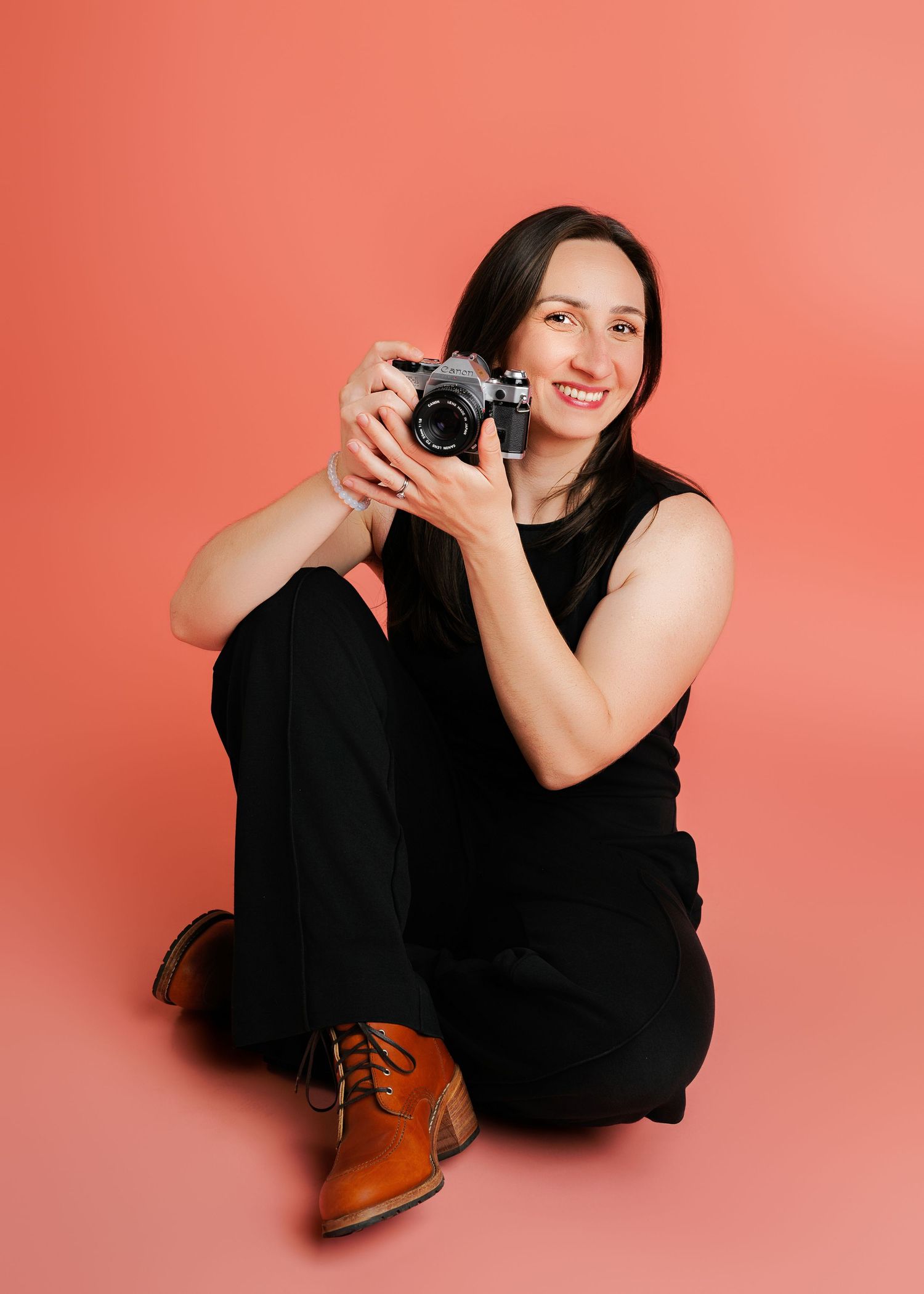 A smiling woman in a black jumpsuit and brown leather boots holding up a vintage camera against a coral pink background.