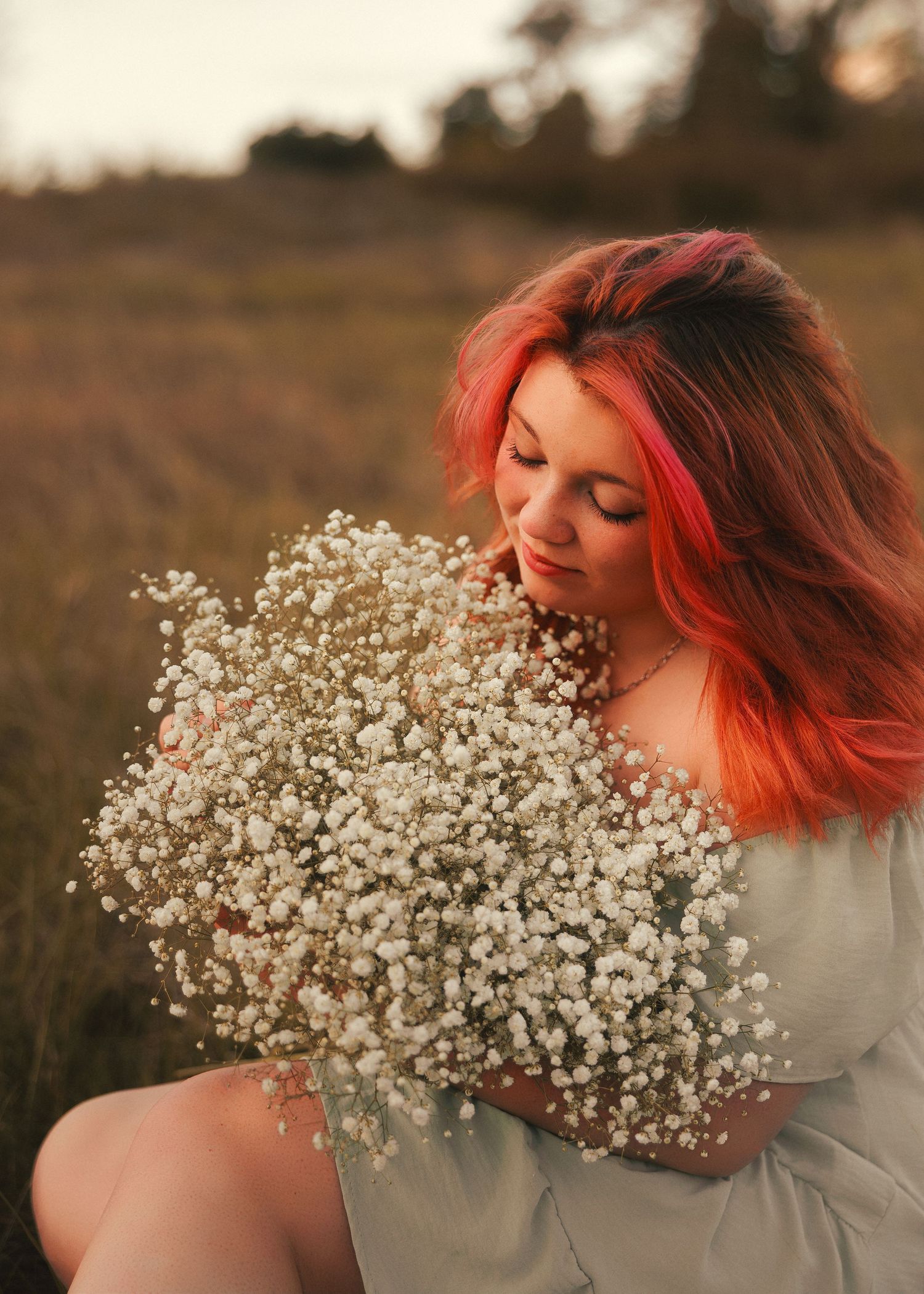 A dreamy portrait in a field at sunset shows someone with coral-colored hair holding a large bouquet of baby's breath flowers.