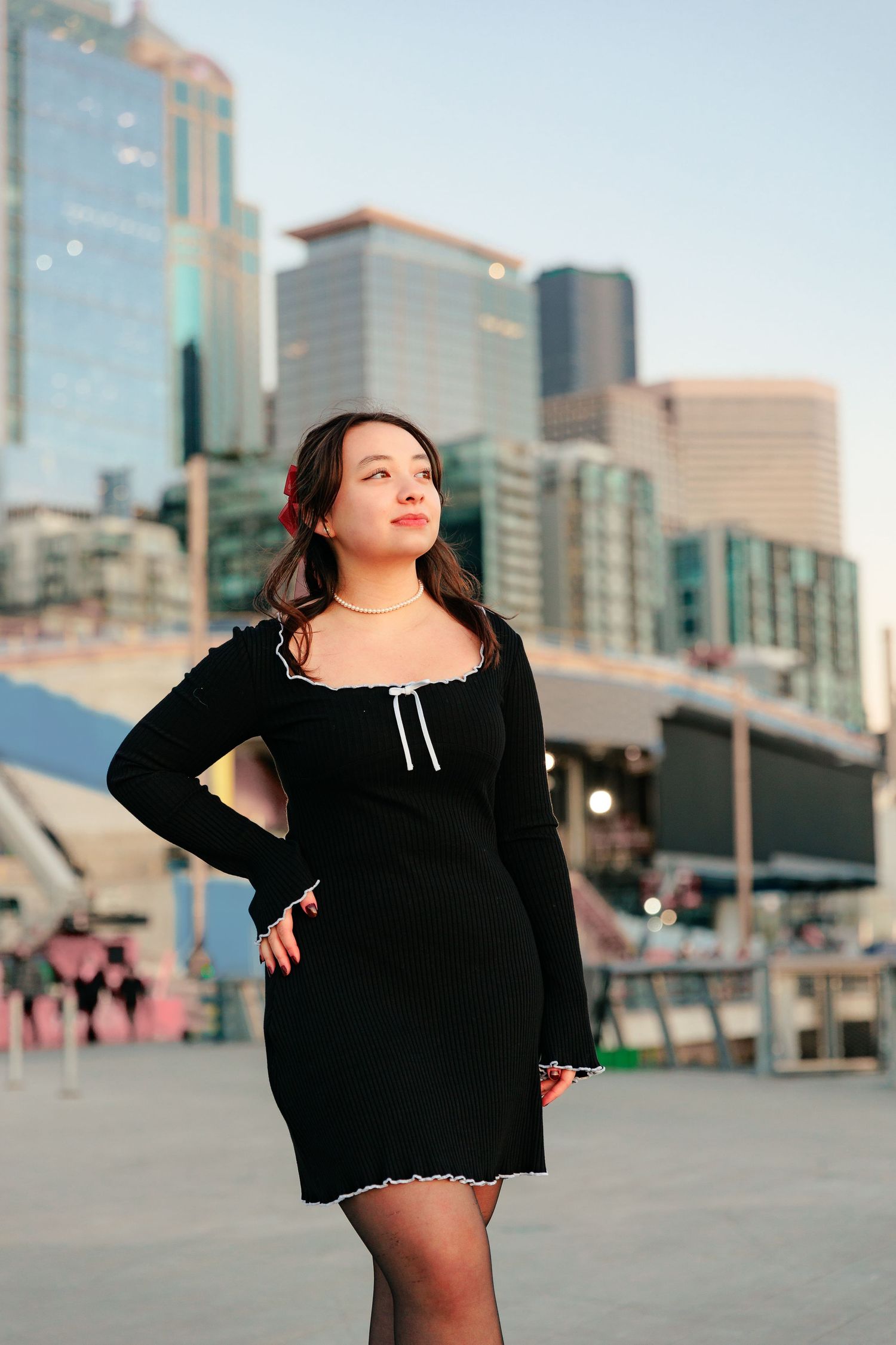 A woman in a black dress poses confidently against a modern city skyline backdrop during golden hour.