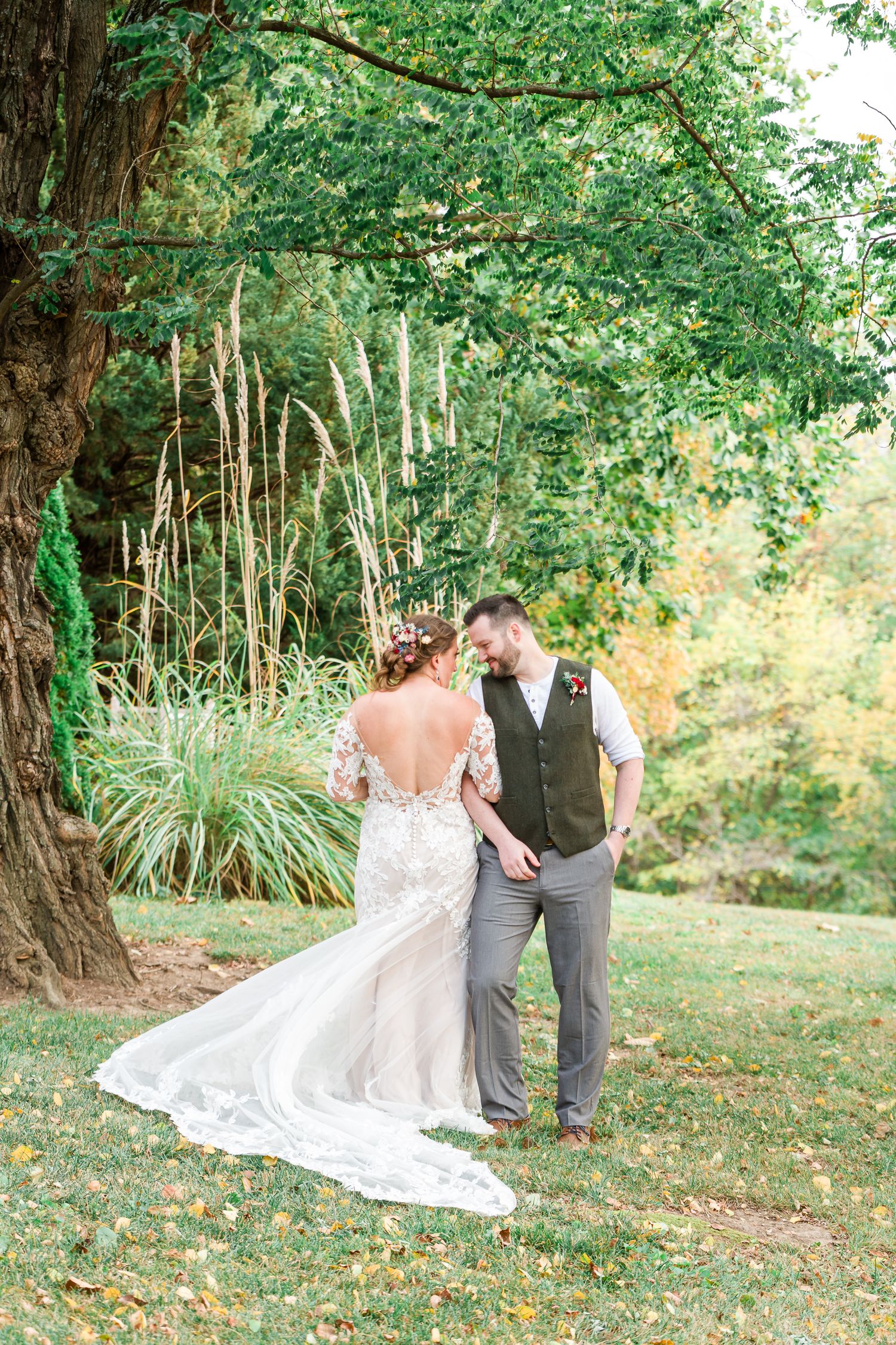 Bride in lace back wedding dress poses with groom under a large tree in a garden setting by Stephanie Coulter Photography