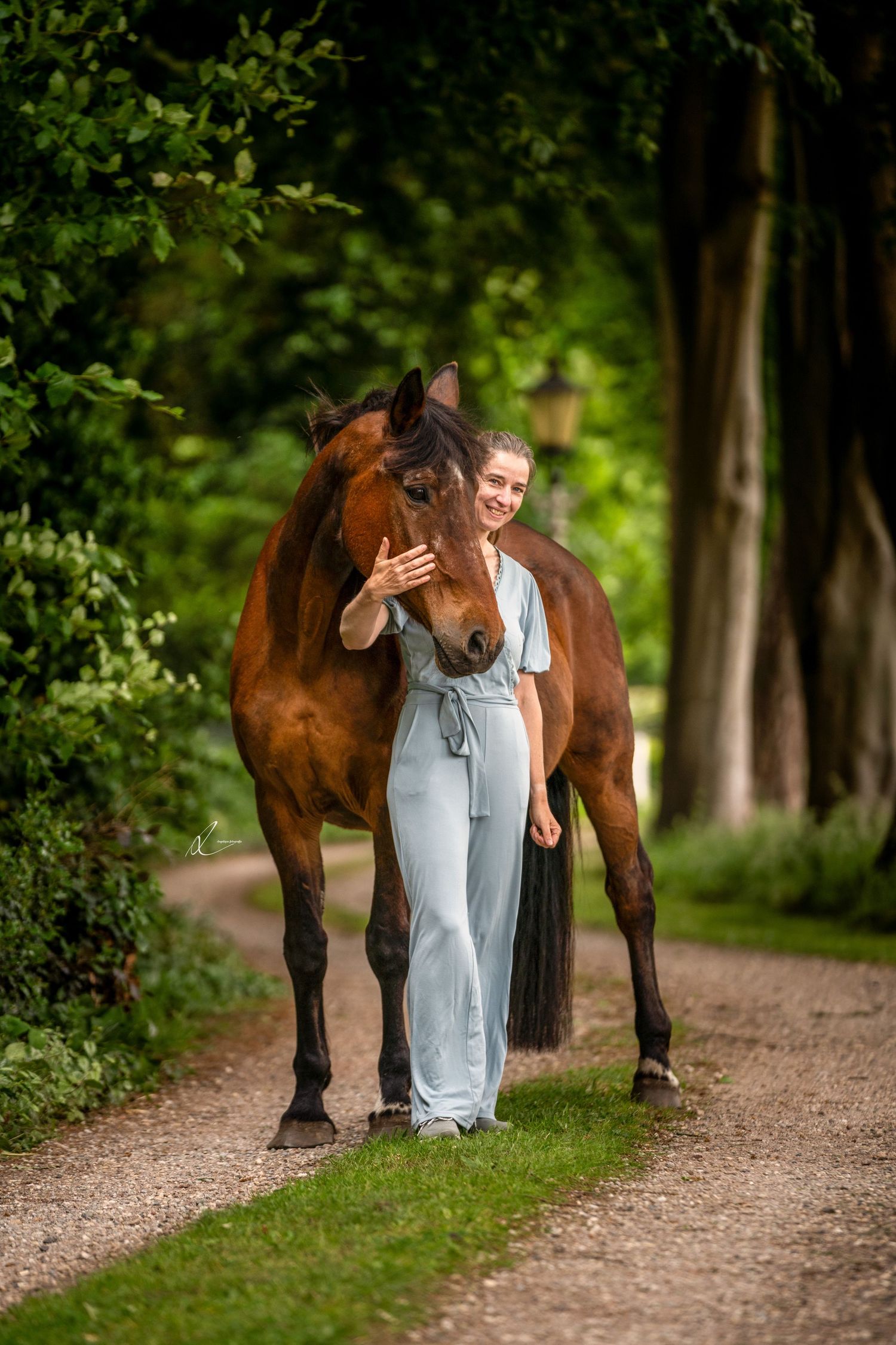 A person in overalls walking along a wooded path shares a tender moment with their brown horse.
