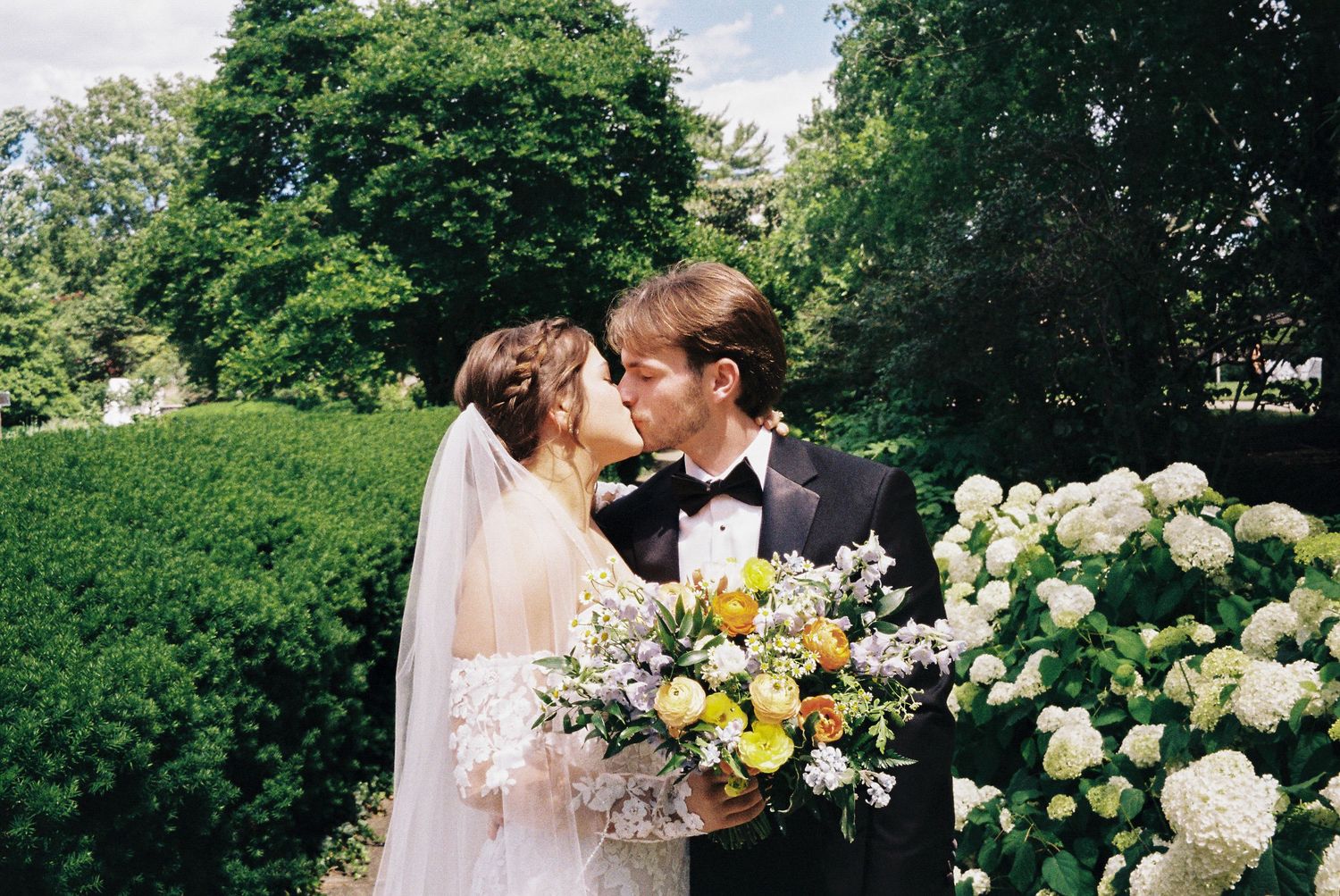 A couple in wedding attire share a romantic moment surrounded by lush gardens and blooming white roses.