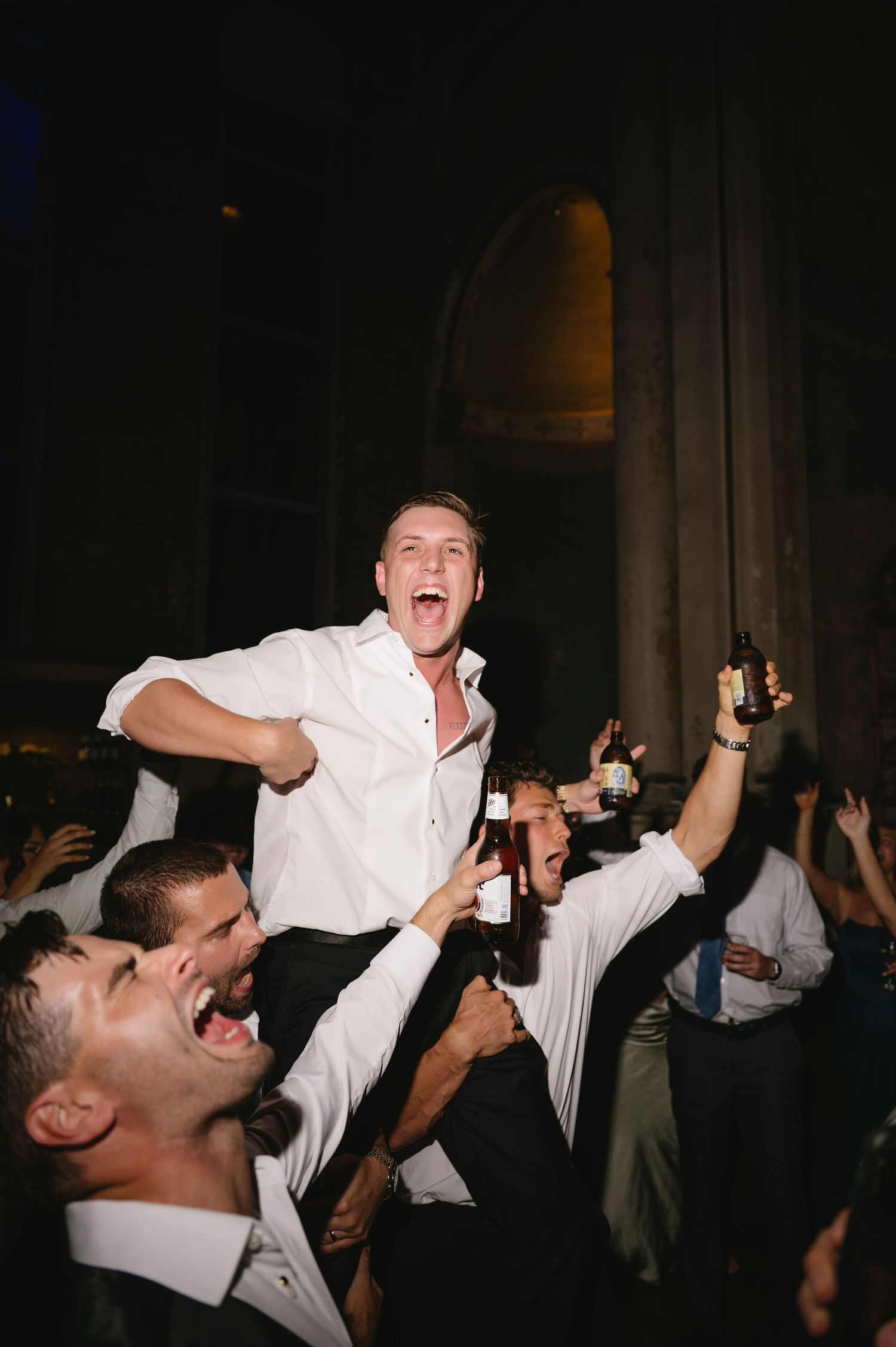 Excited party guests in white shirts raise their drinks and cheer during a lively celebration in a dark venue.