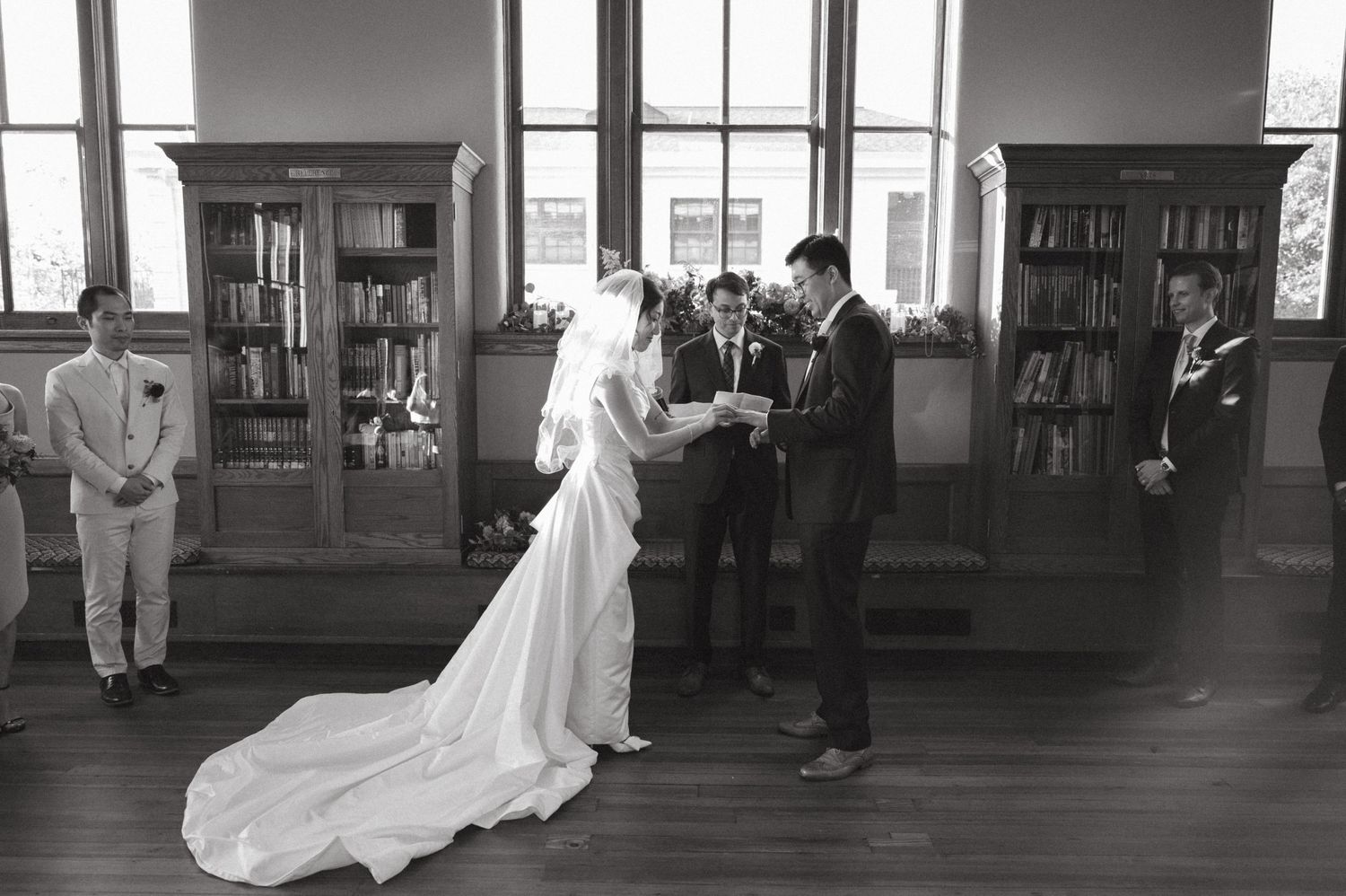 A wedding ceremony takes place in a library setting with wooden floors and built-in bookshelves.