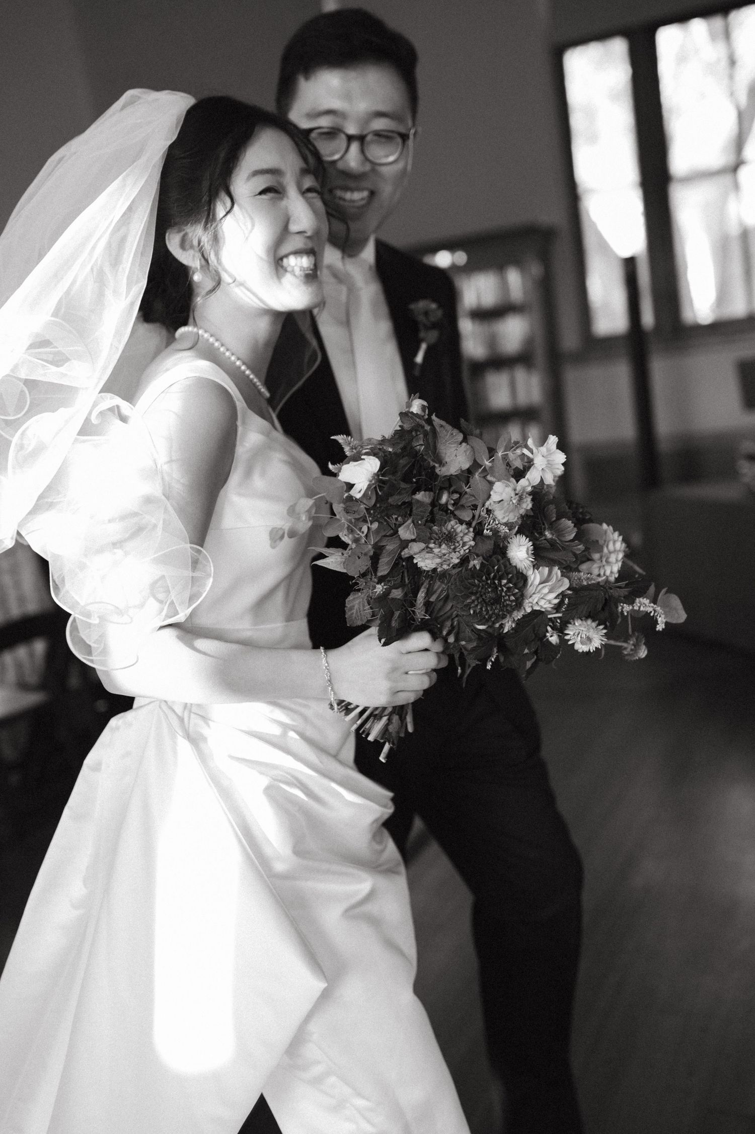 A joyful black and white wedding photo showing a bride in white dress holding flowers with her partner laughing together.