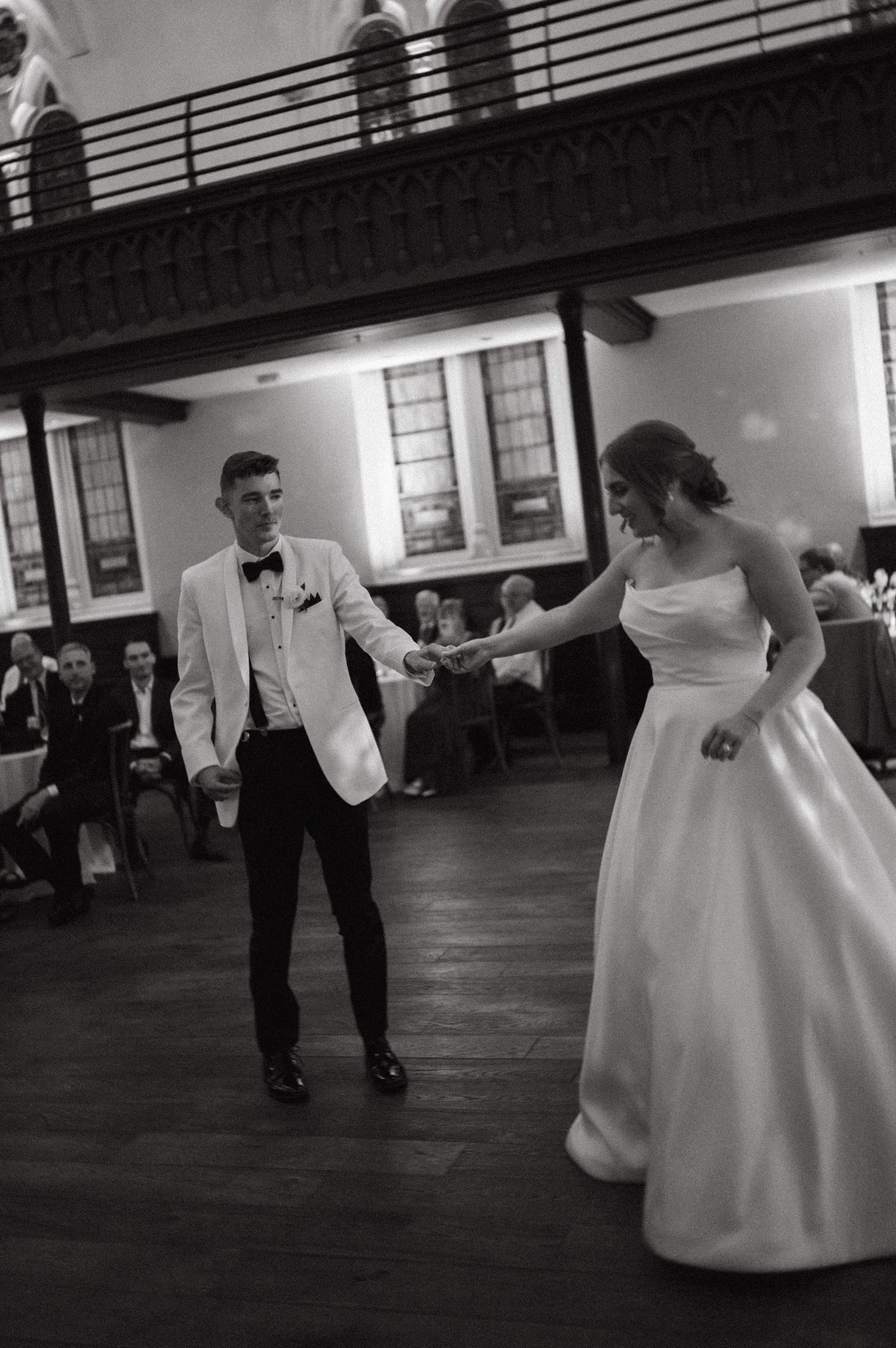 A couple shares their first dance in black and white on a wooden ballroom floor.