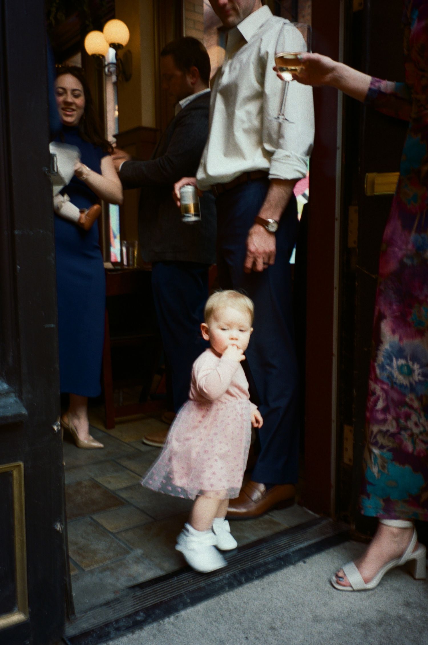 A small child in a pink dress takes their first steps across a doorway threshold while adults watch nearby.