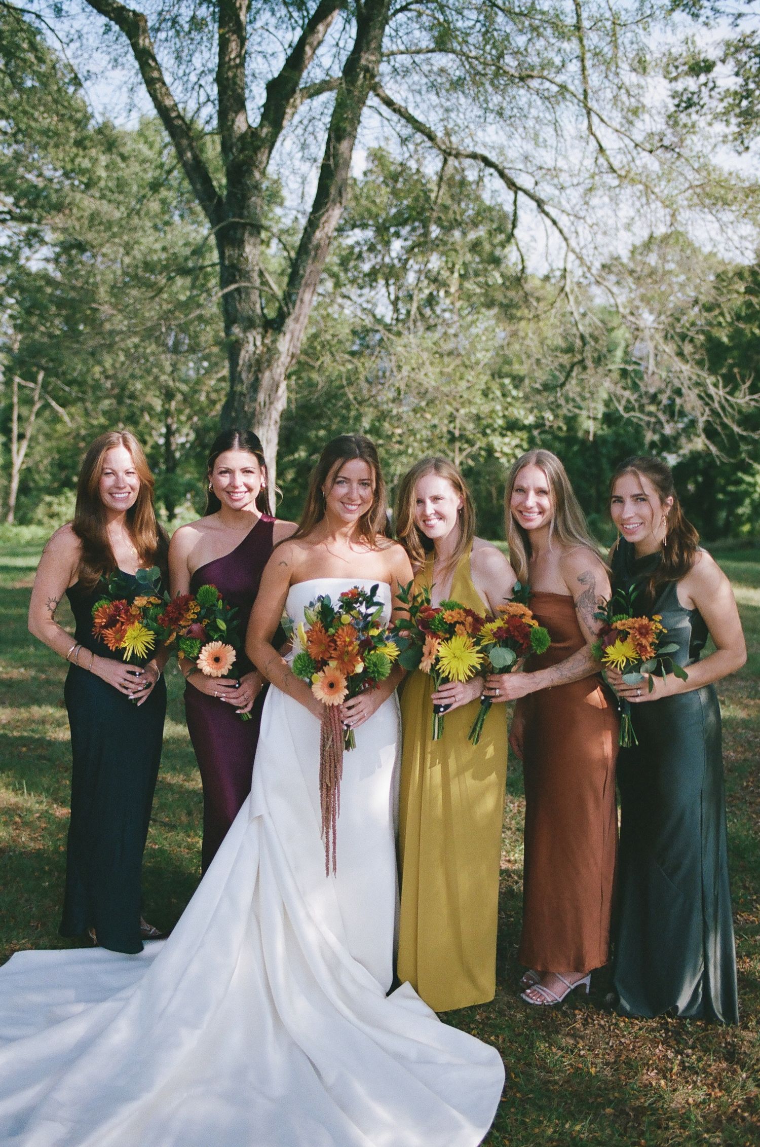 A bridal party in autumn colors poses outdoors with sunflowers and fall bouquets among trees in a park setting.