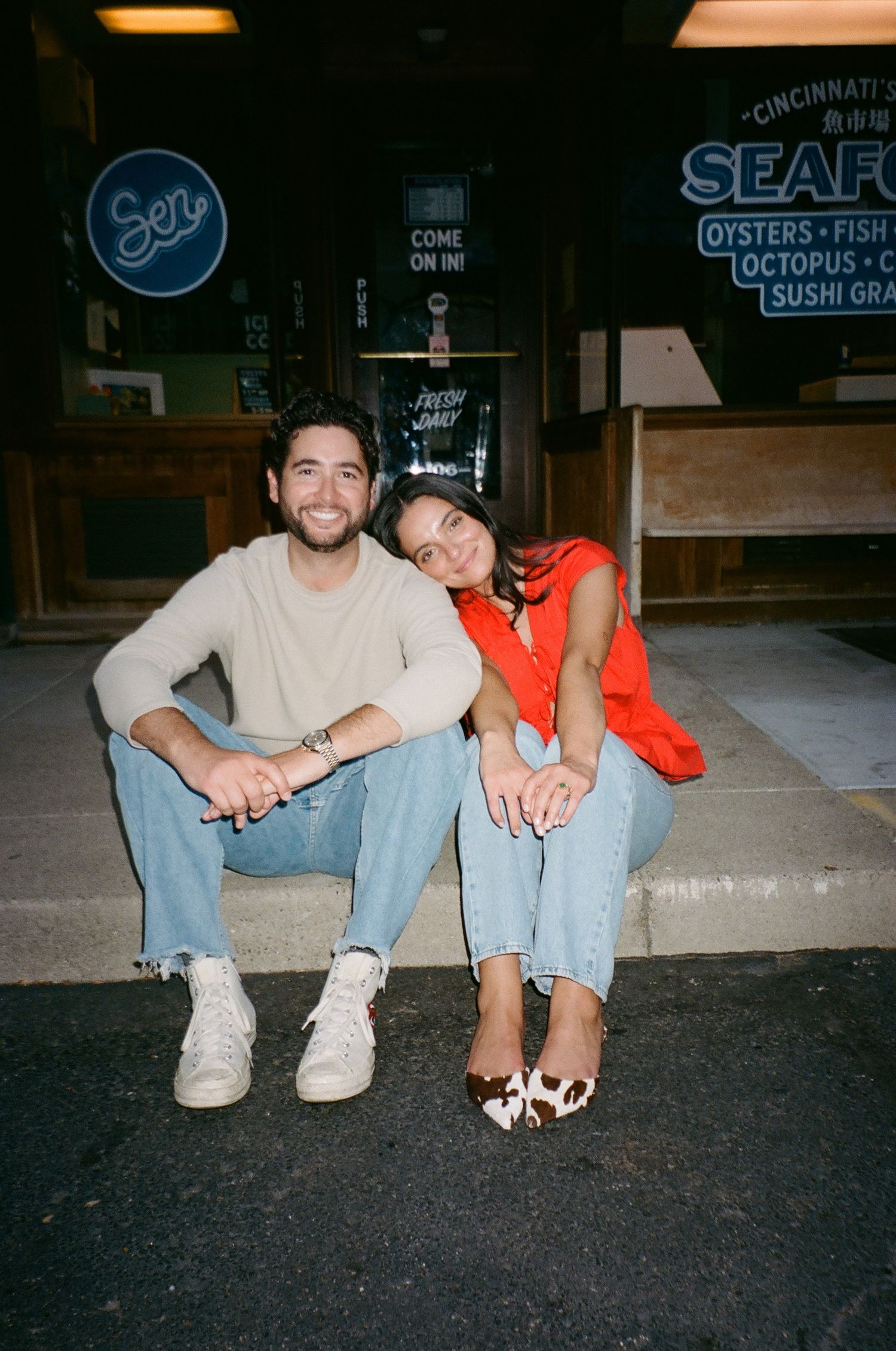 Two people sit casually on a curb at night wearing jeans and sneakers, shown in a series of candid shots.
