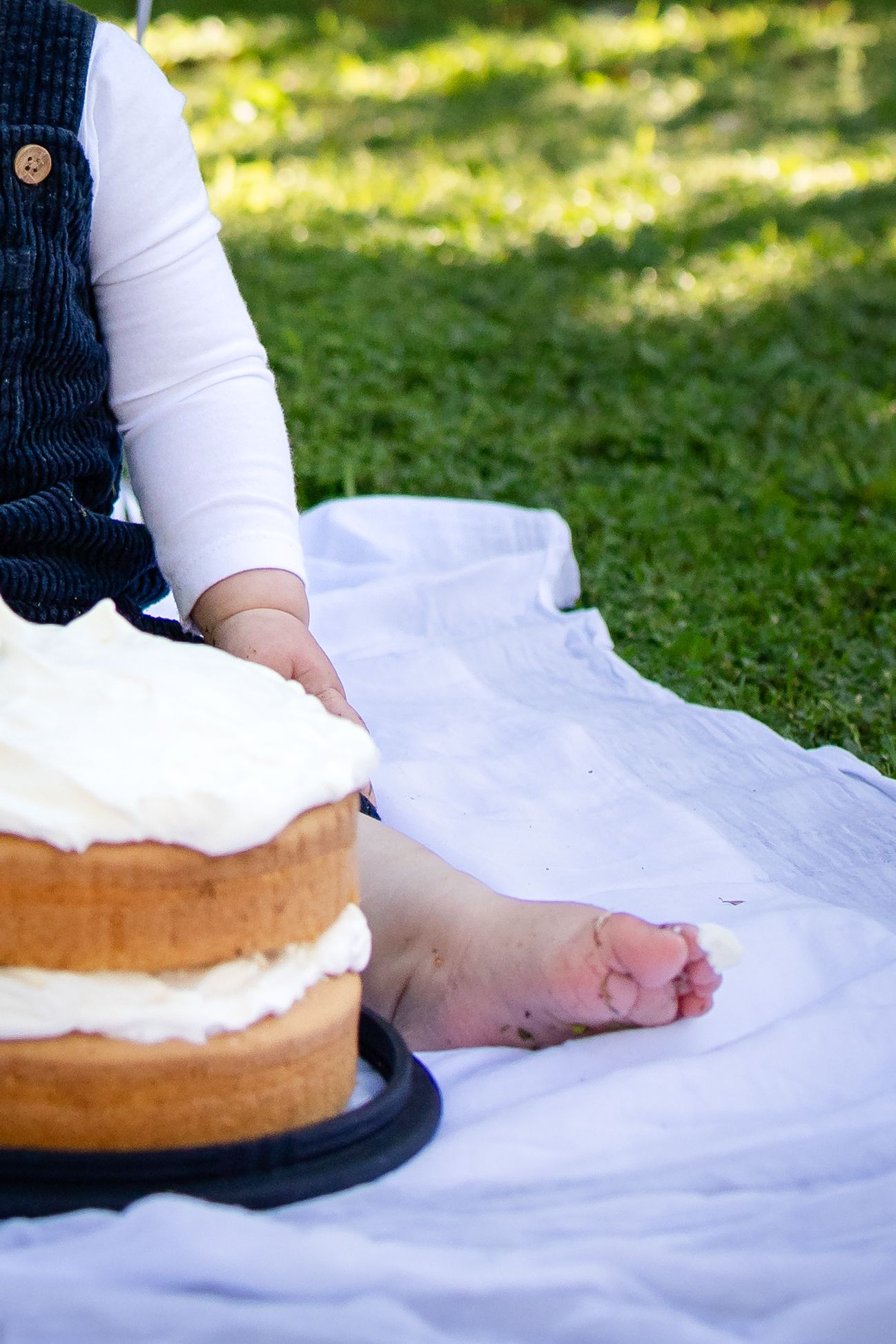 Baby sitting on white blanket in grass beside a two-layer white frosted cake on a black stand.