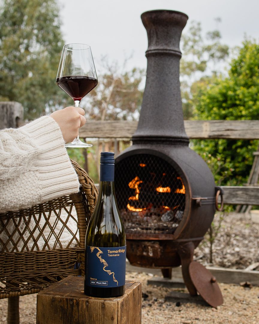 Person holding red wine glass beside a TarraWarra Estate Sangiovese bottle near a lit cast iron chiminea outdoors.