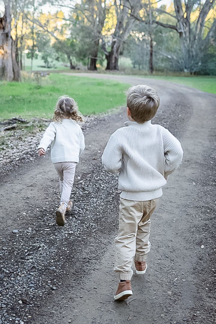 Two young children in cream sweaters and tan pants run along a gravel path surrounded by tall trees.