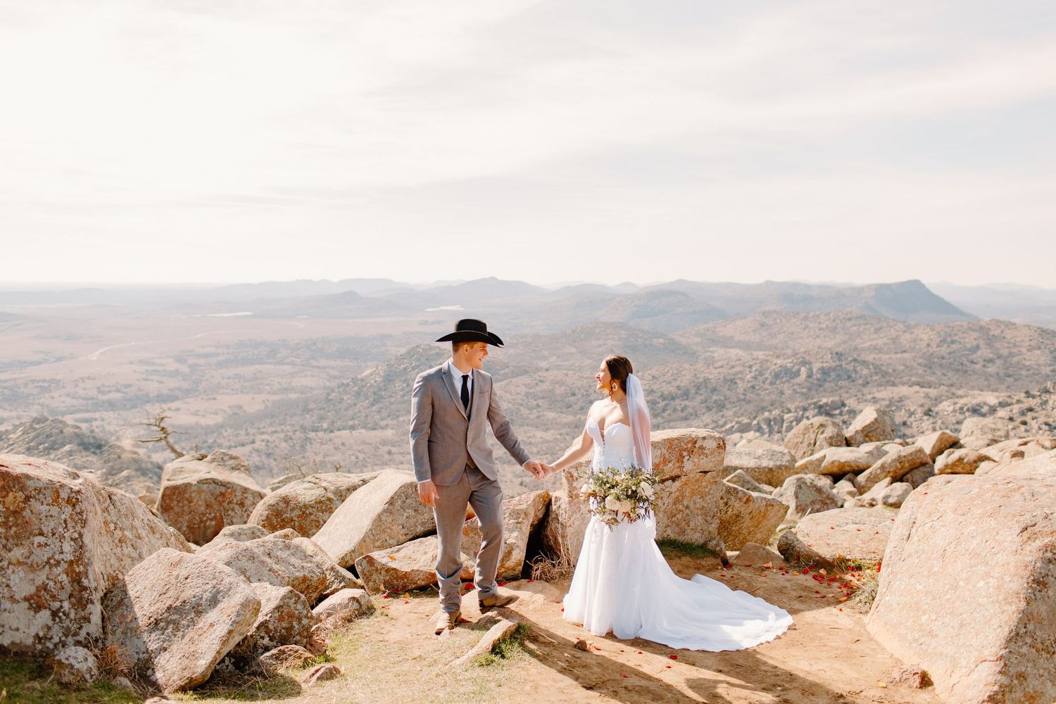 A bride and groom hold hands while walking among rocky terrain with a vast mountain landscape.