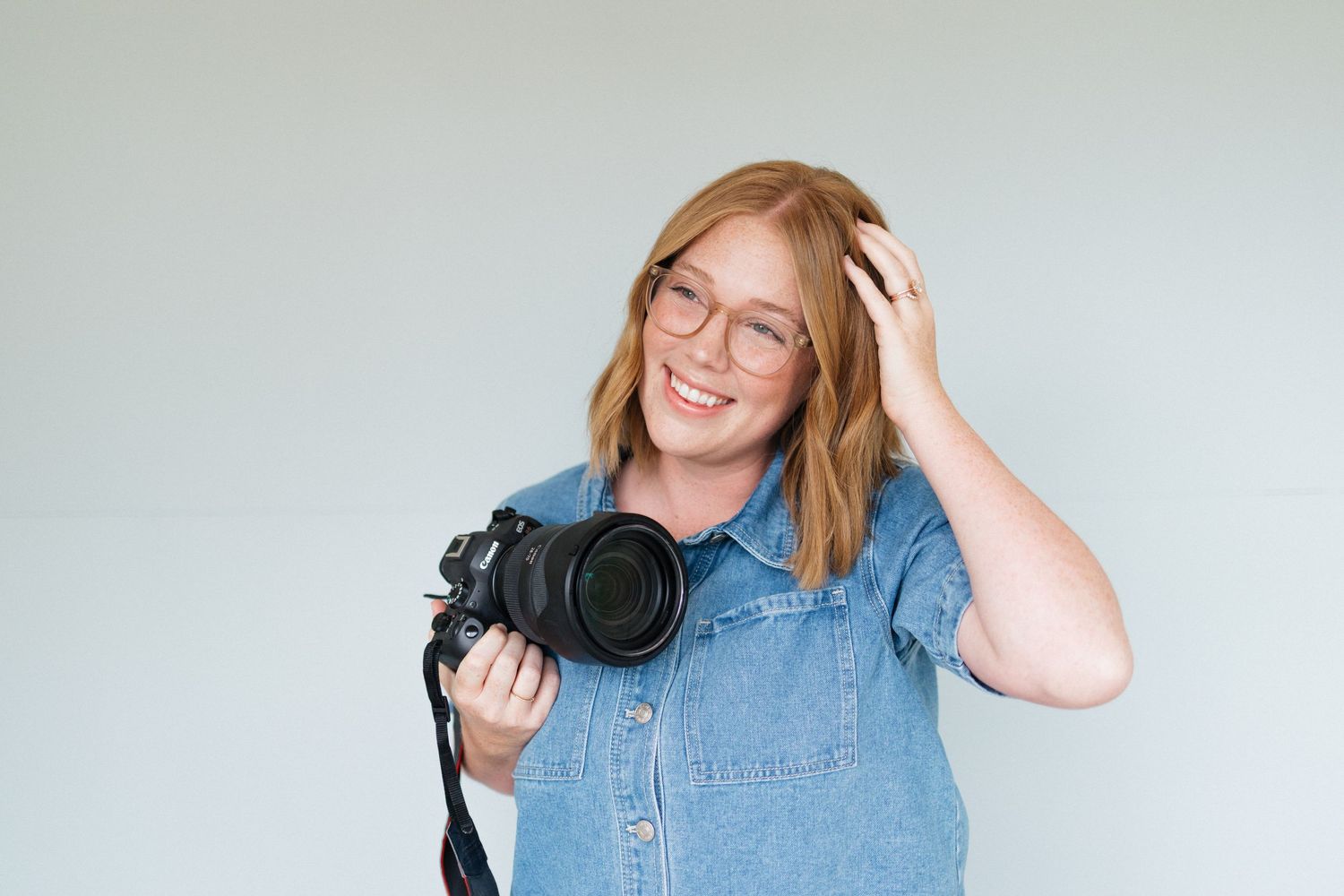A smiling female photographer with glasses holding a camera and touching her hair on gray background.