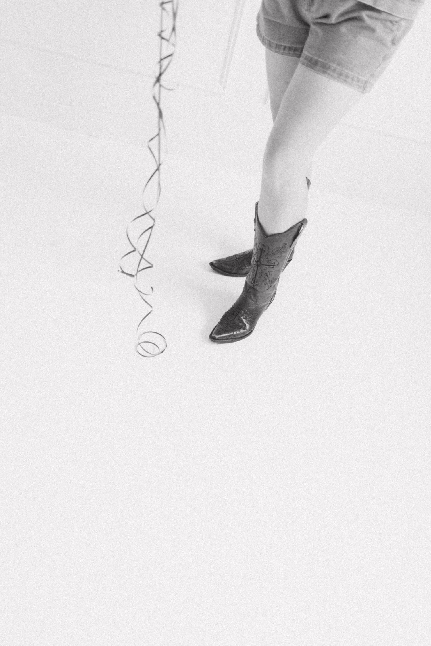Black and white photo of a person wearing cowboy boots holding balloon ribbons on a white floor.
