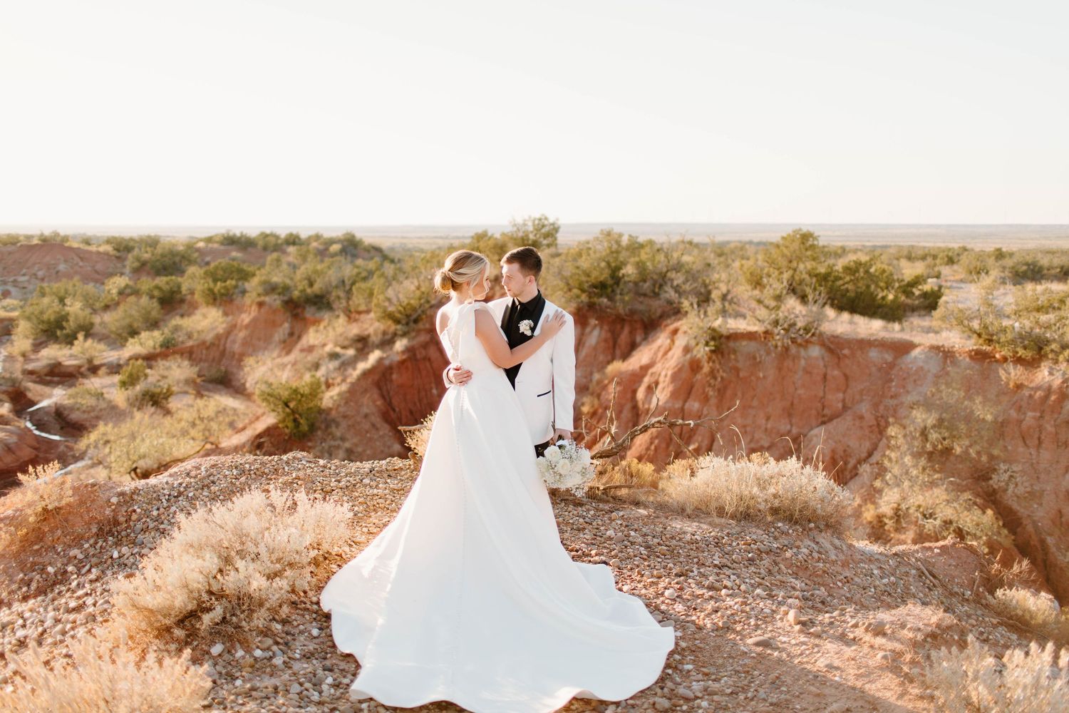 Bride and groom embrace on dramatic desert canyon landscape, her white gown flowing in the warm golden light.