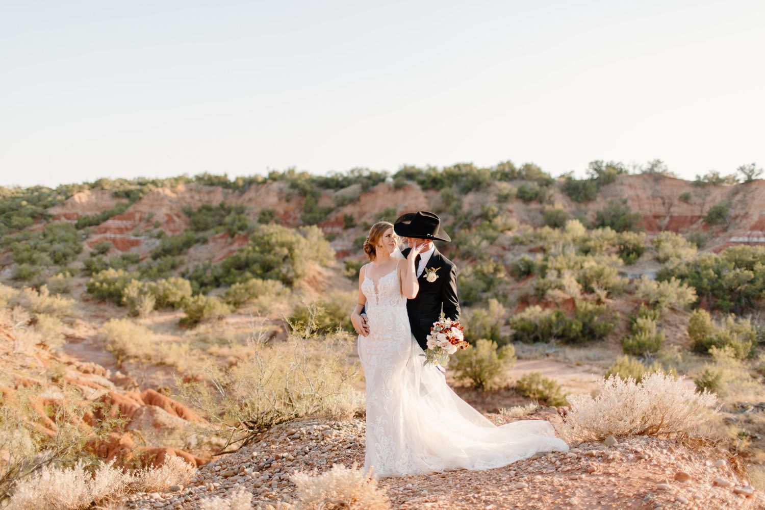 Bride and groom embrace on scenic desert landscape with red rock formations at sunset.