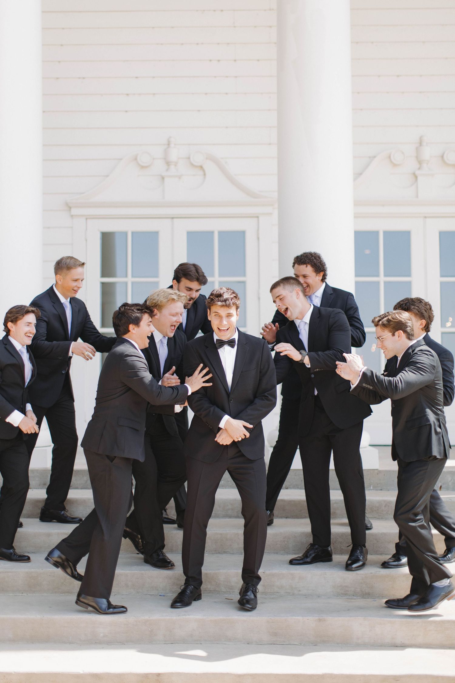 Groom in black tuxedo surrounded by laughing groomsmen in dark suits on steps of a white columned wedding venue.