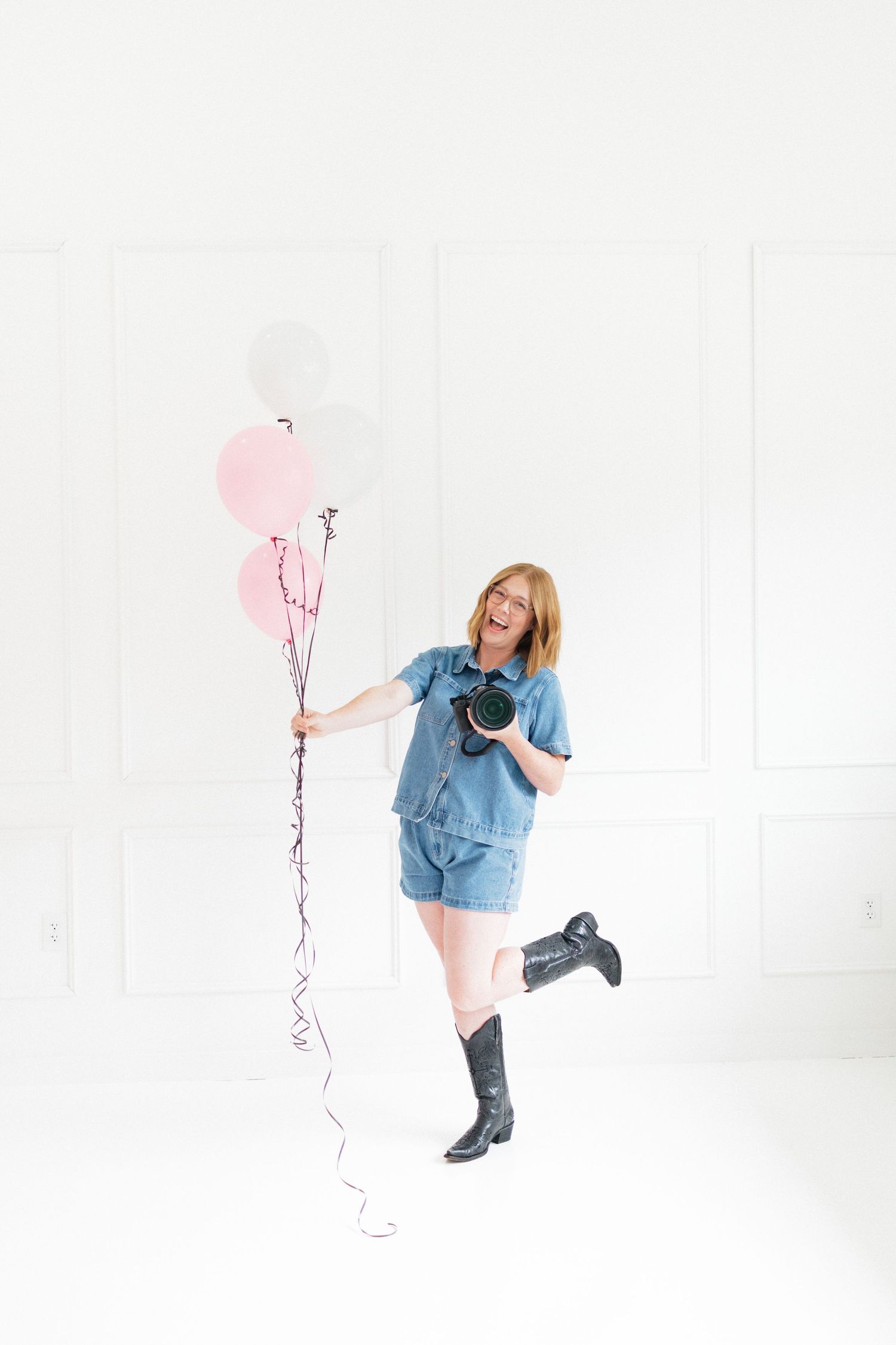 A cheerful redhead woman in denim holds pink balloons and a camera in a bright white room.