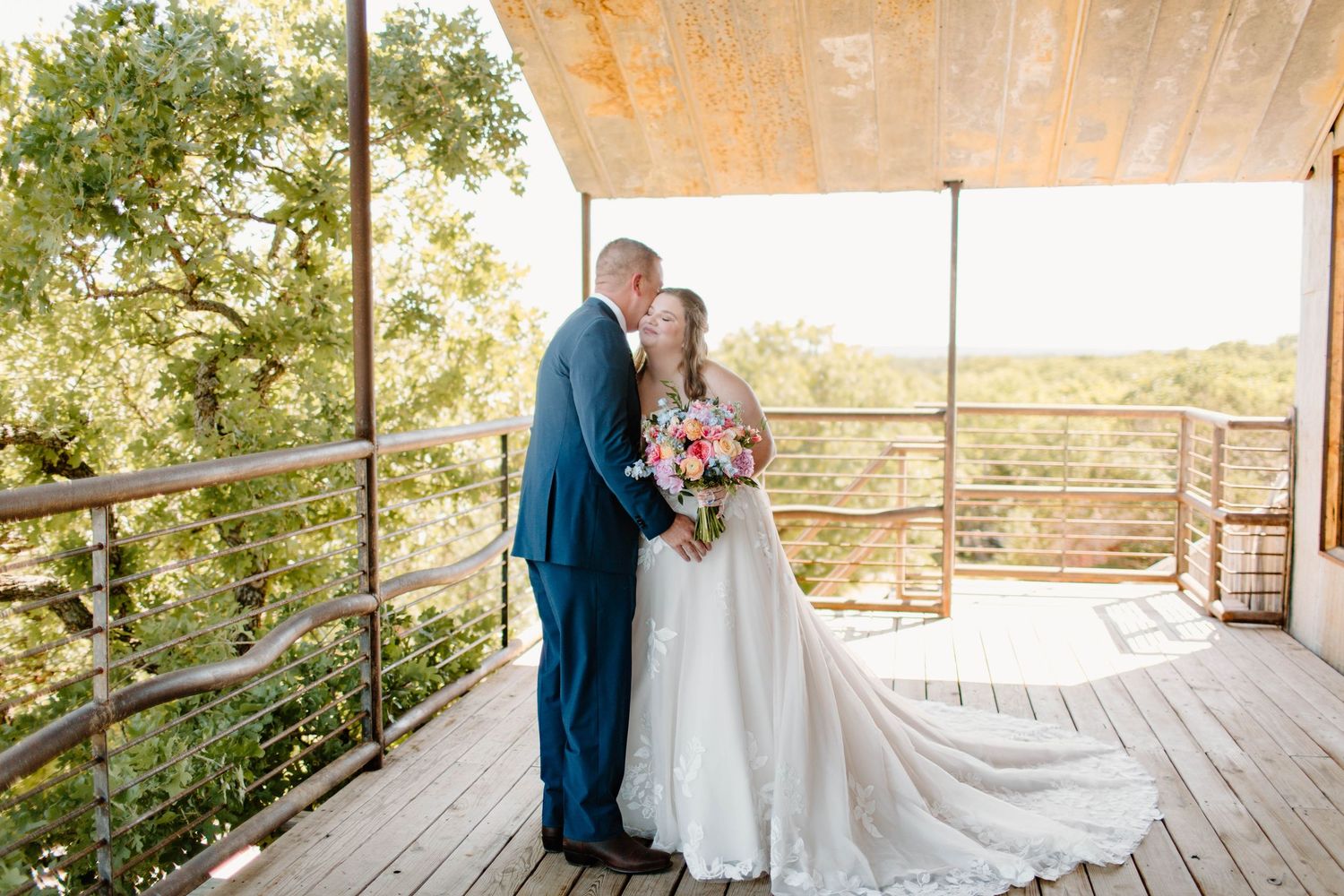 Bride and groom share a tender kiss on a wooden deck overlooking lush green trees at an outdoor wedding venue.