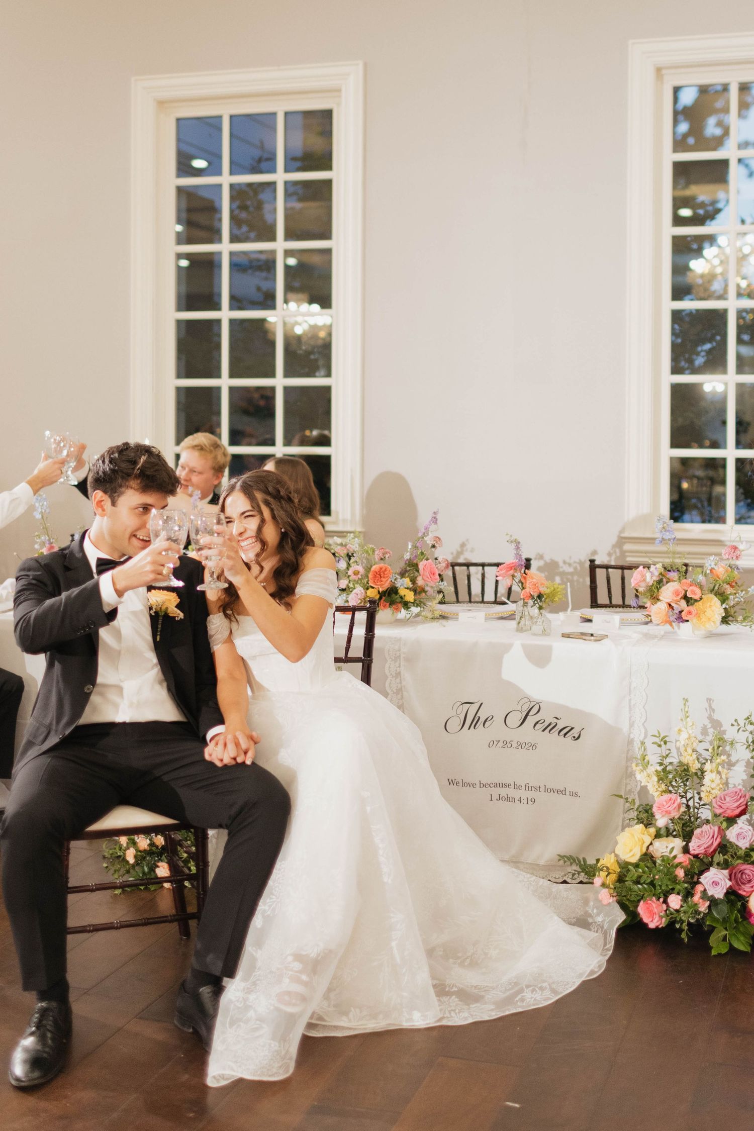 Bride and groom toast champagne at sweetheart table with floral arrangements at reception.